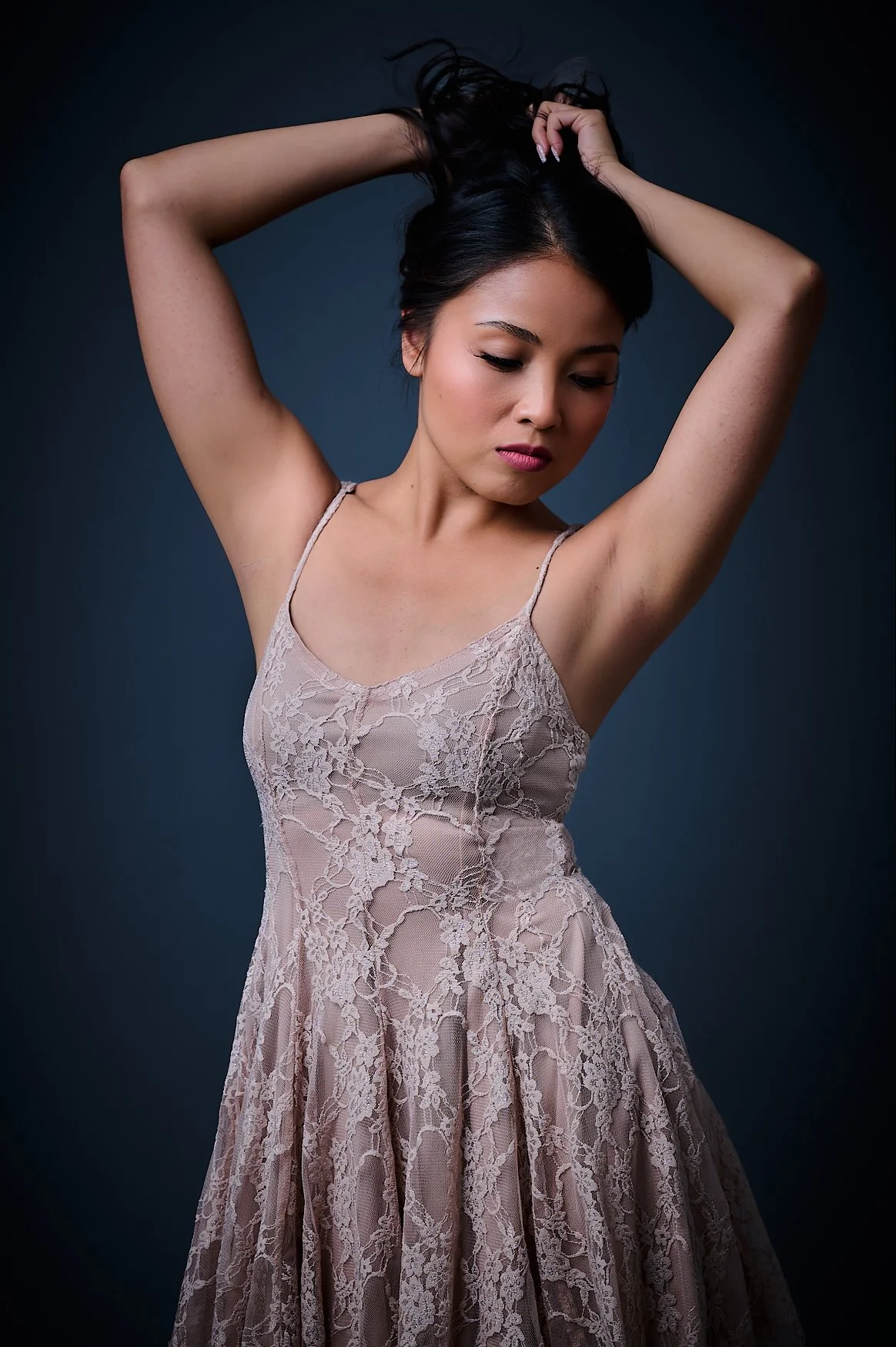 A woman with black hair in a lace dress is posing with her arms raised and hands in her hair against a dark background.