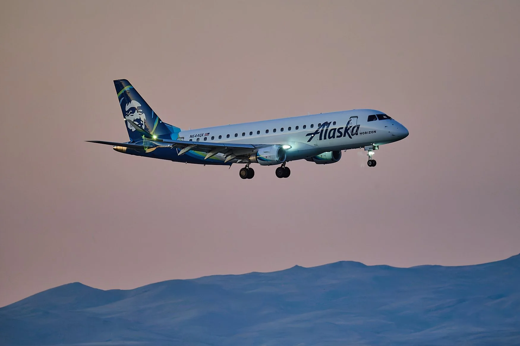 An Alaska Airlines airplane flying in the sky with mountains below, during sunset or dusk.