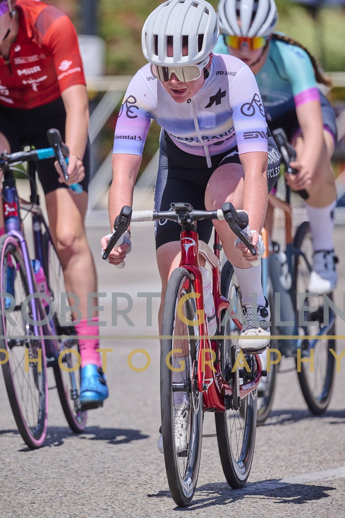 Female cyclist wearing a white helmet and sunglasses competing in a bike race with other female cyclists on a sunny day.