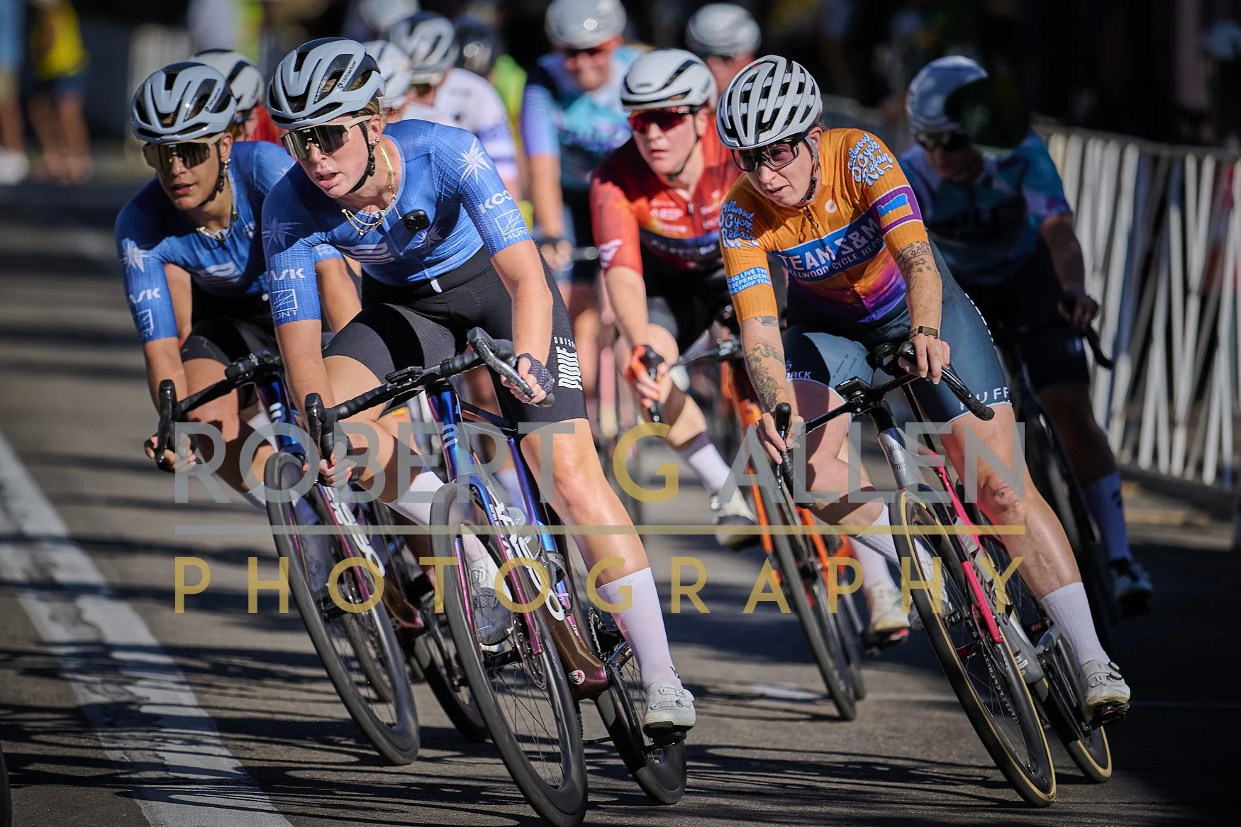 Group of female cyclists in a race, wearing helmets and sunglasses, riding closely together on a city street.