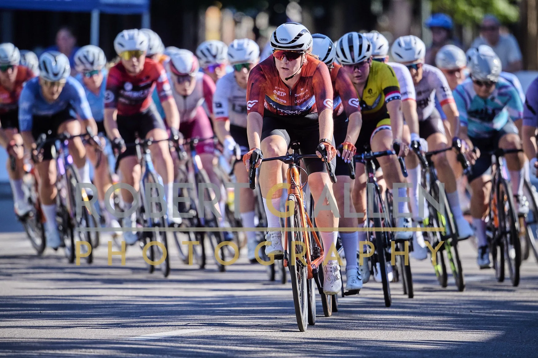 Group of competitive female cyclists riding on a road during a race, wearing helmets and colorful jerseys.