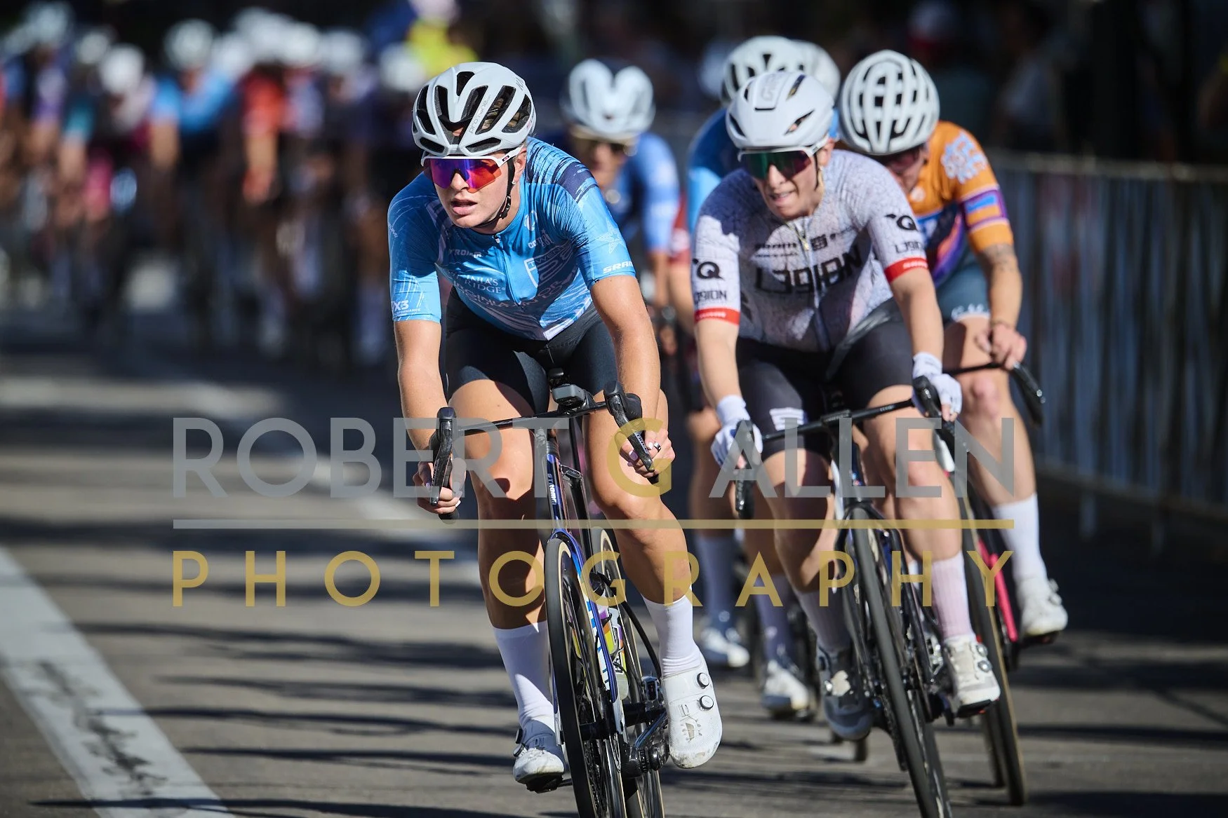 Cyclists participating in a race, wearing helmets and colorful jerseys, riding on a paved road with a crowd of spectators behind metal barricades.