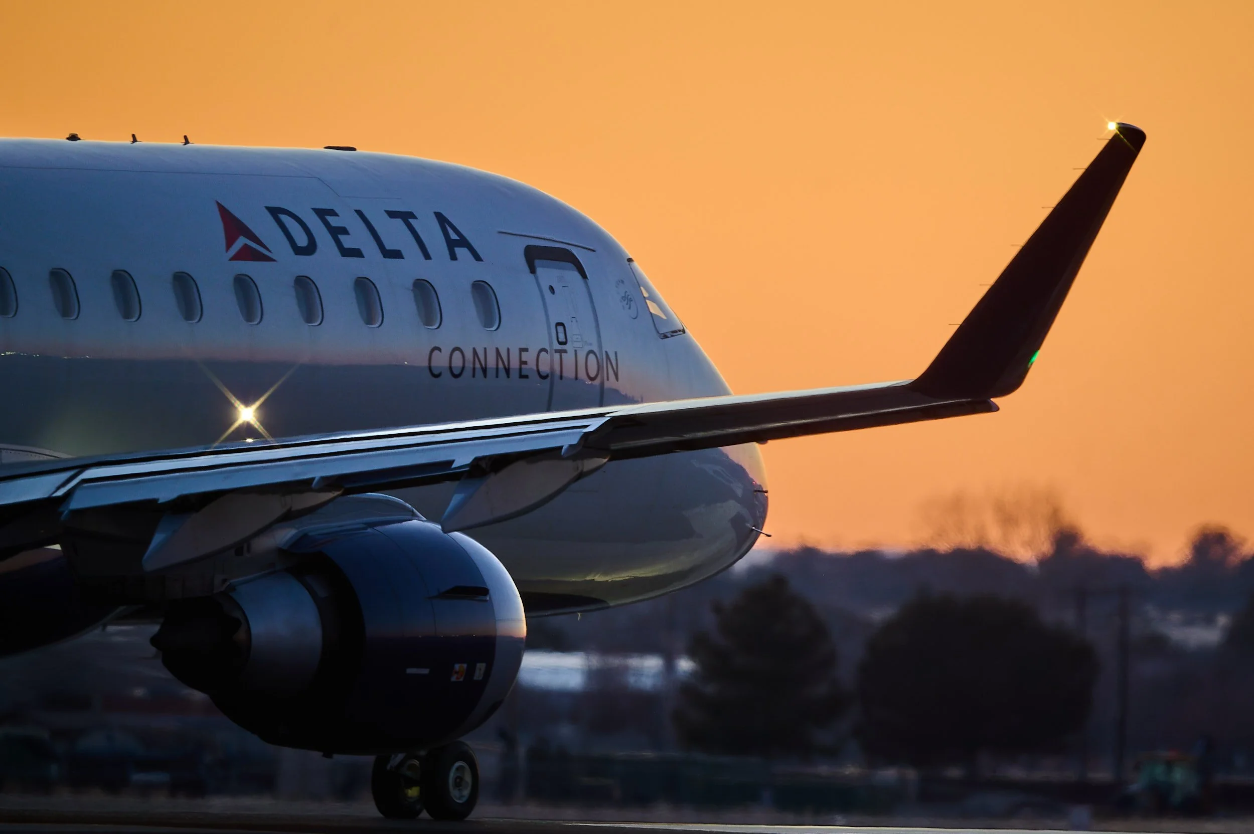 Front view of a Delta airplane at sunset, showing the nose, cockpit windows, engine, wingtip, and some ground features.