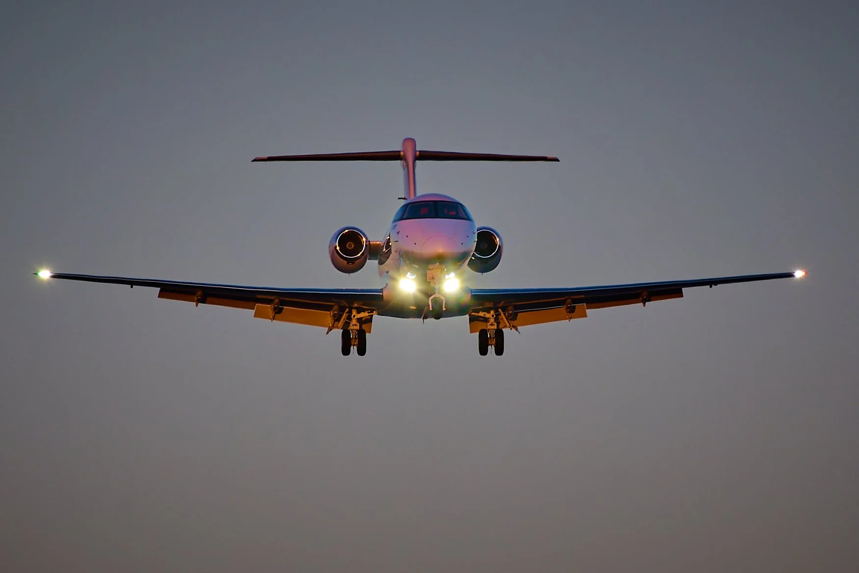 Front view of a commercial airplane flying towards the camera at dusk, with its lights illuminated.