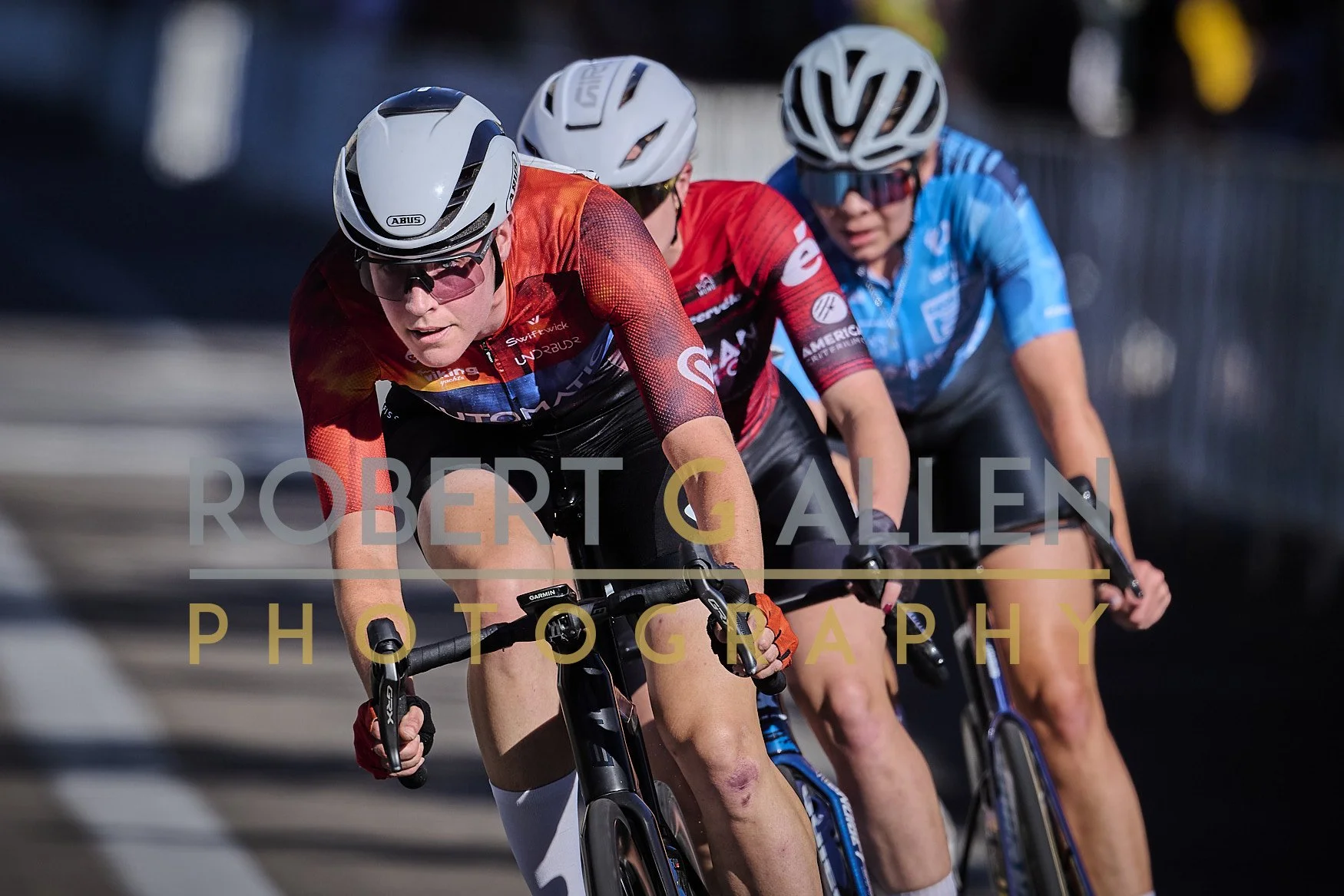 Three cyclists racing on a road, wearing helmets and athletic gear, with a focus on the lead rider.