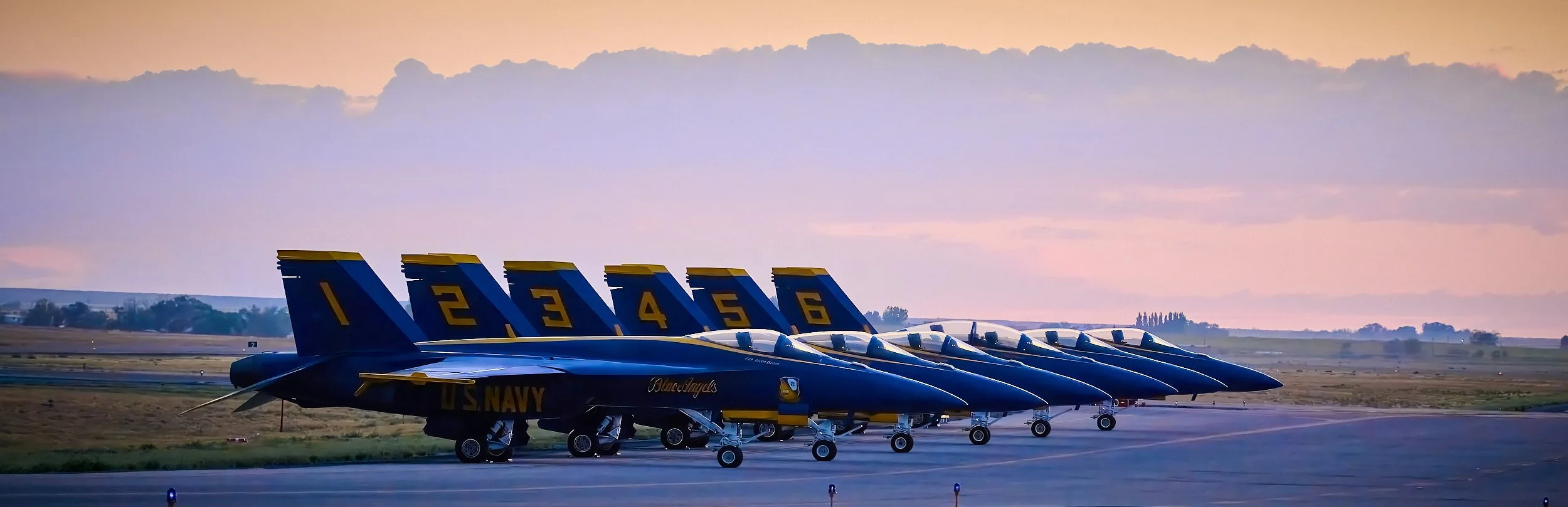 A lineup of six U.S. Navy Blue Angels fighter jets parked on an airport runway during sunset, with colorful sky and distant horizon.