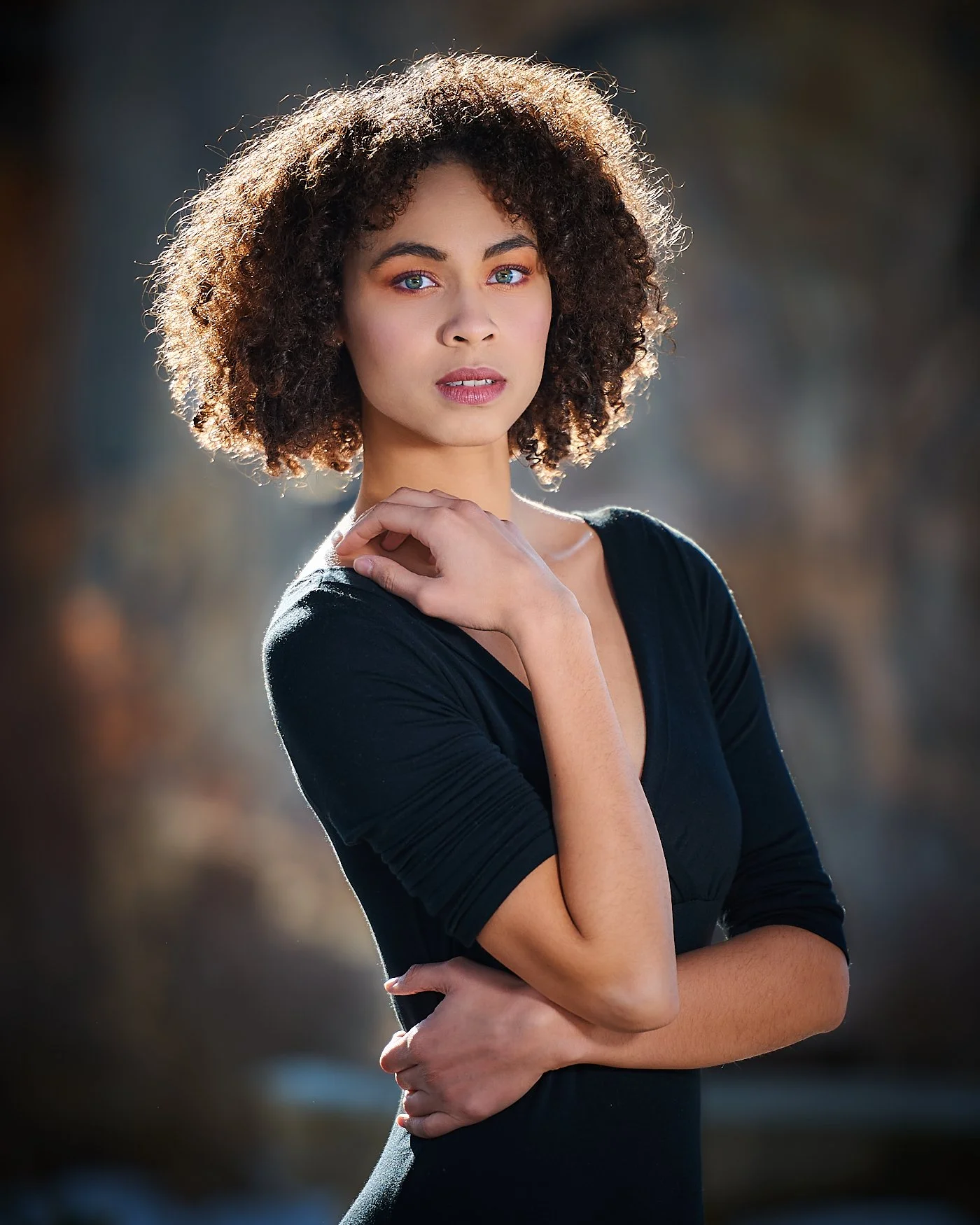 Portrait of a young woman with curly hair, wearing a black top, standing outdoors with natural sunset lighting.