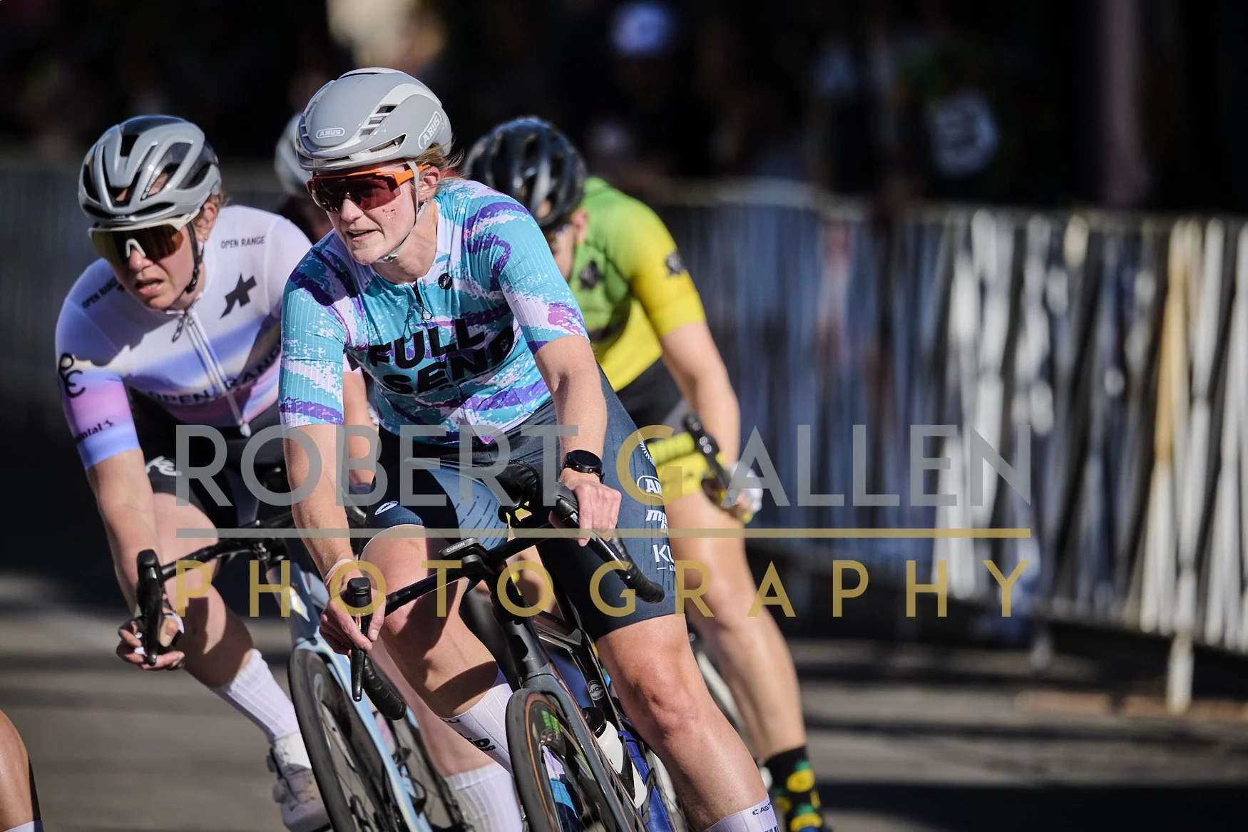 Cyclists racing on a velodrome track, wearing helmets and sports gear, with a focus on the lead cyclist in a blue jersey.