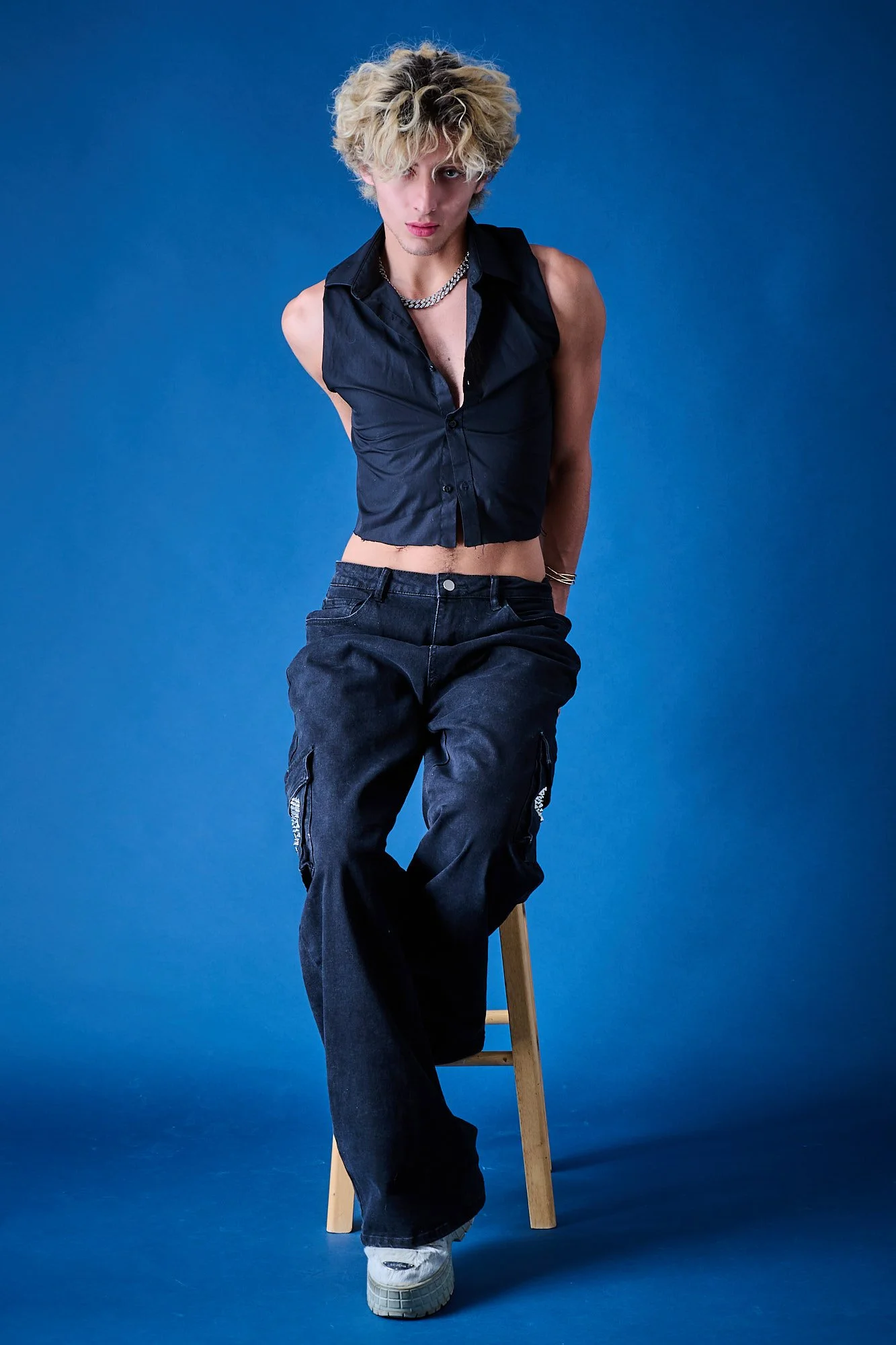 Young man with curly blonde hair sitting on a wooden stool against a blue background. Wearing a partially unbuttoned black sleeveless shirt, a silver chain, dark cargo pants, and sneakers.