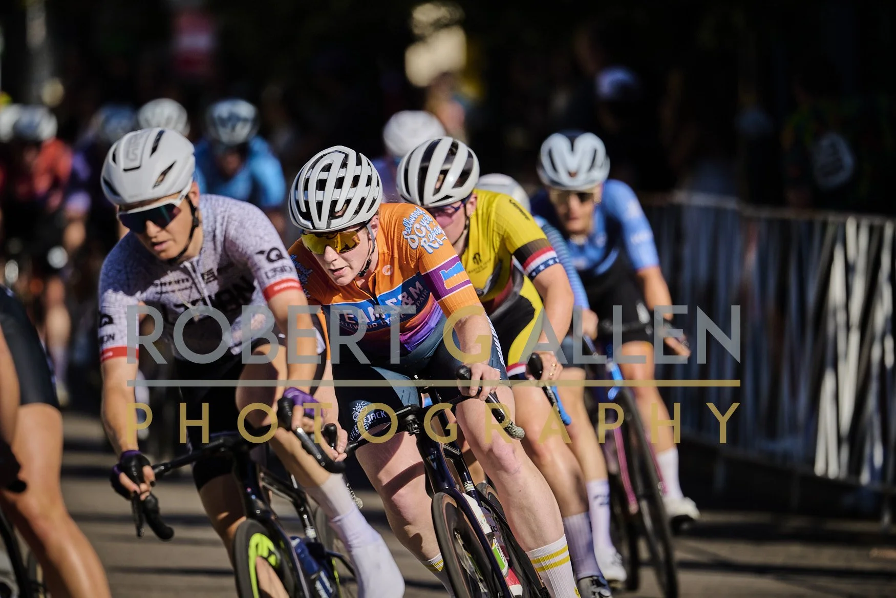 Cyclists competing in a race, wearing helmets and colorful jerseys. They are focused and riding closely together on a sunny day.