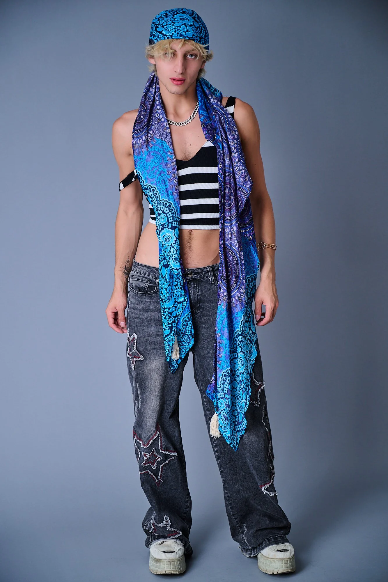 A young man with blonde curly hair, wearing a blue patterned bandana, a black and white striped crop top, a silver chain necklace, distressed baggy jeans with patches and star designs, white sneakers, and a blue paisley scarf draped around his neck, 