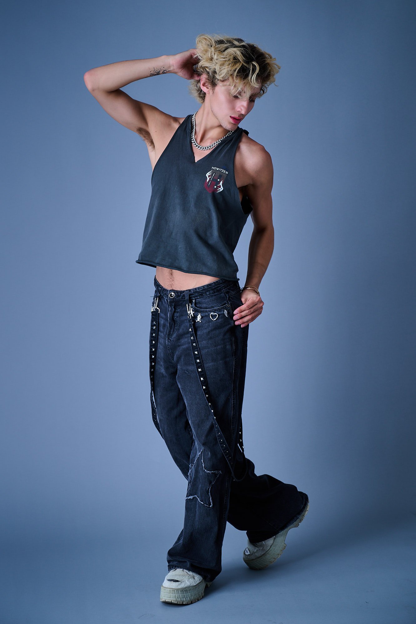 A young person with blonde, curly hair and light skin, wearing a sleeveless black top with graphic logo, baggy dark jeans with chains, and chunky sneakers, standing against a plain gray background with one arm raised behind their head.