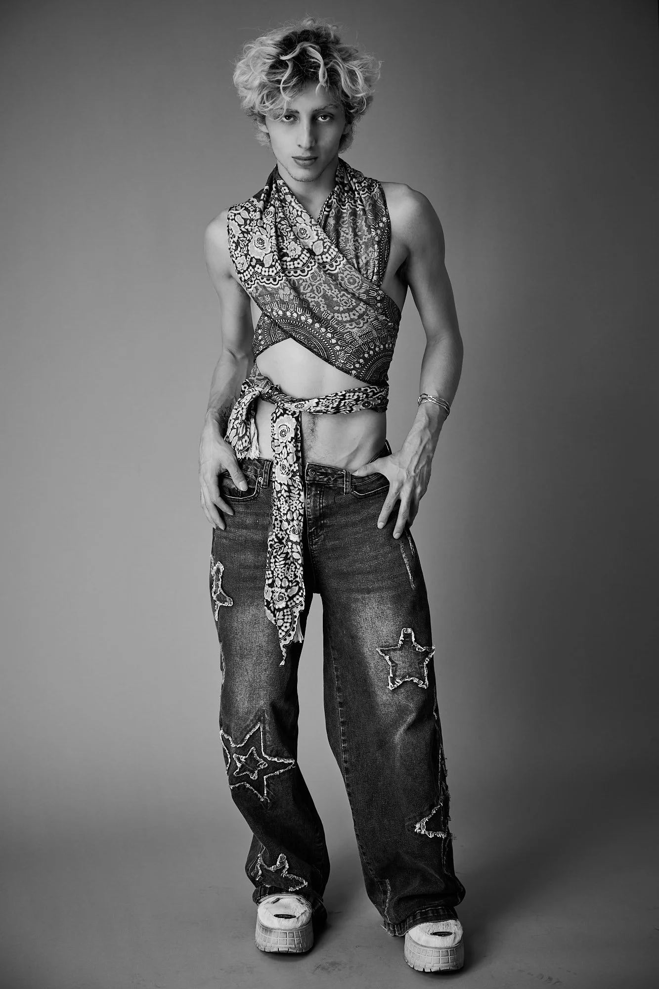 Black and white photo of androgynous person with curly hair wearing patterned scarf, ripped jeans with star patches, and sneakers, posing against a plain background.