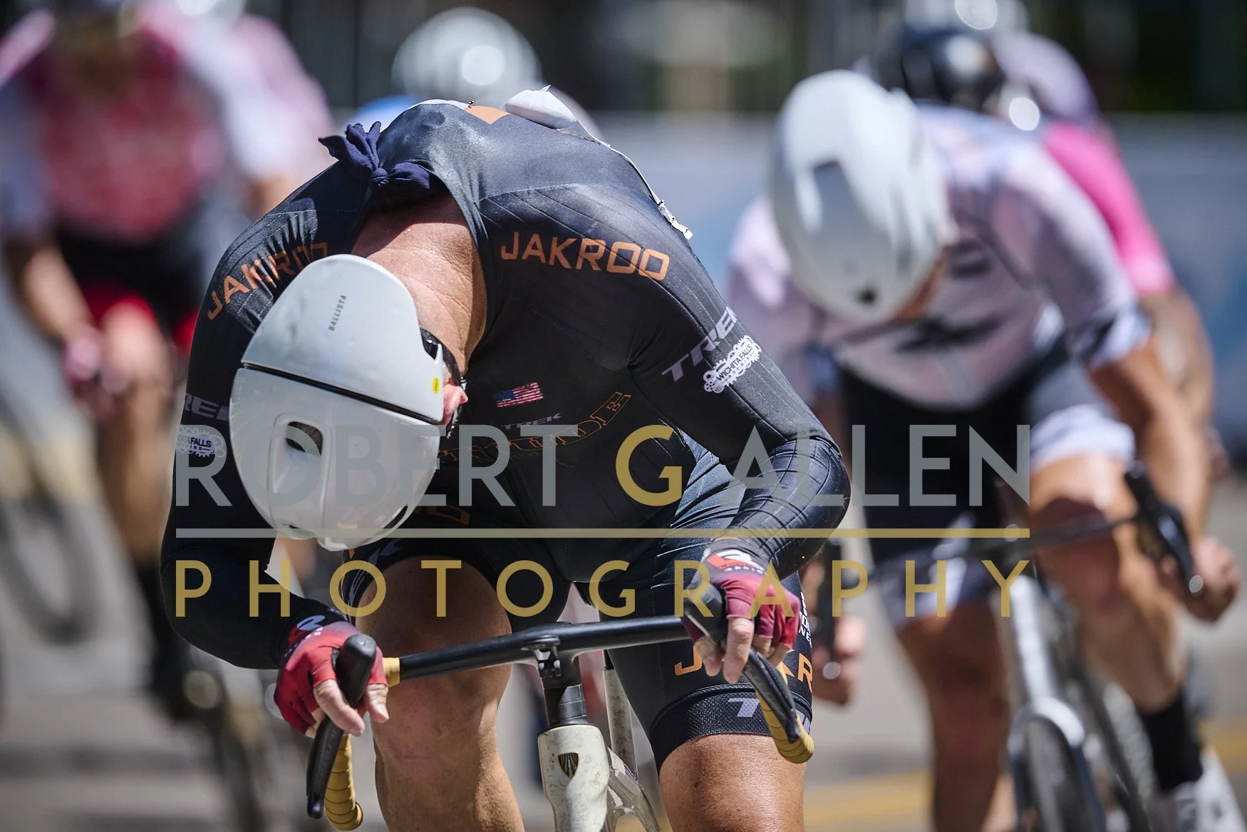 Cyclists wearing helmets and racing gear on bikes during a race, with one cyclist in front leaning forward and looking downward.