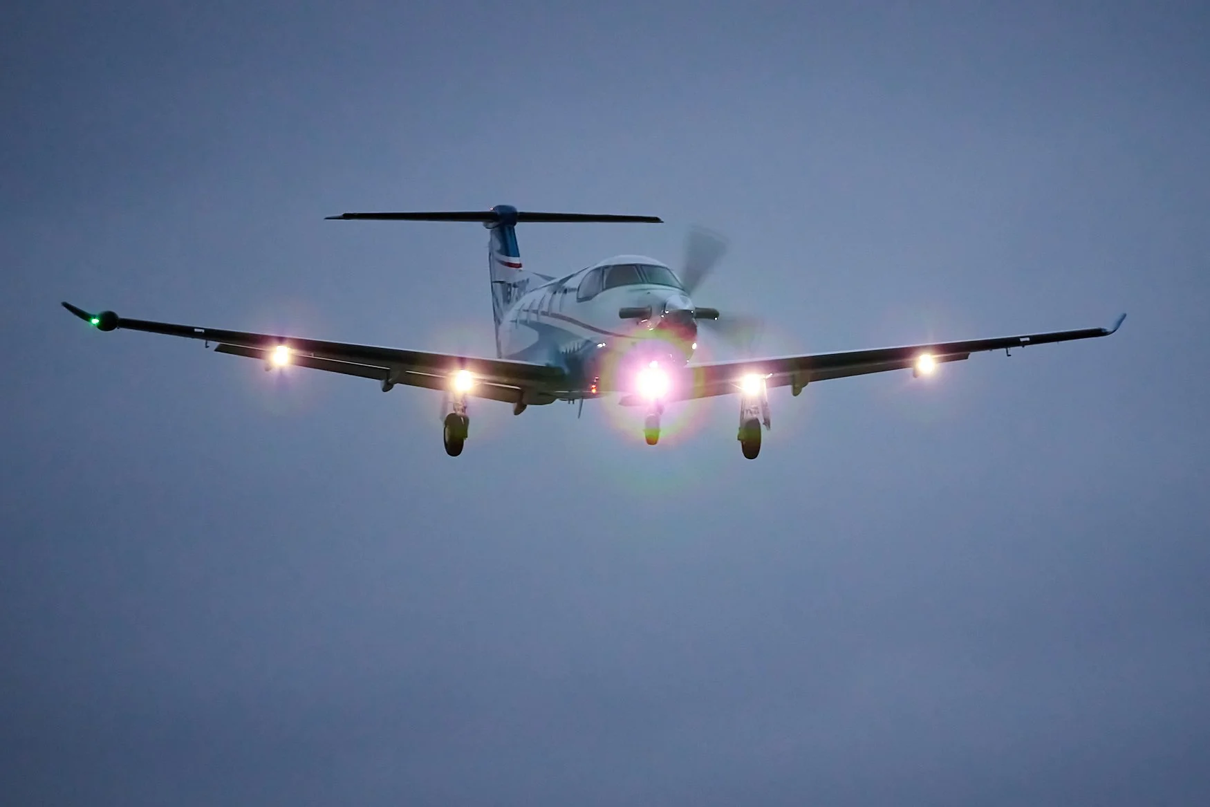 A small airplane flying against a darkening sky with bright landing lights and navigation lights.