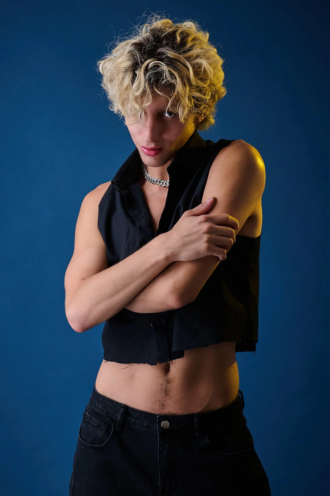 A person with curly blond hair wearing a sleeveless black shirt, a silver chain necklace, and black pants, standing against a dark blue background, with arms crossed and a confident expression.