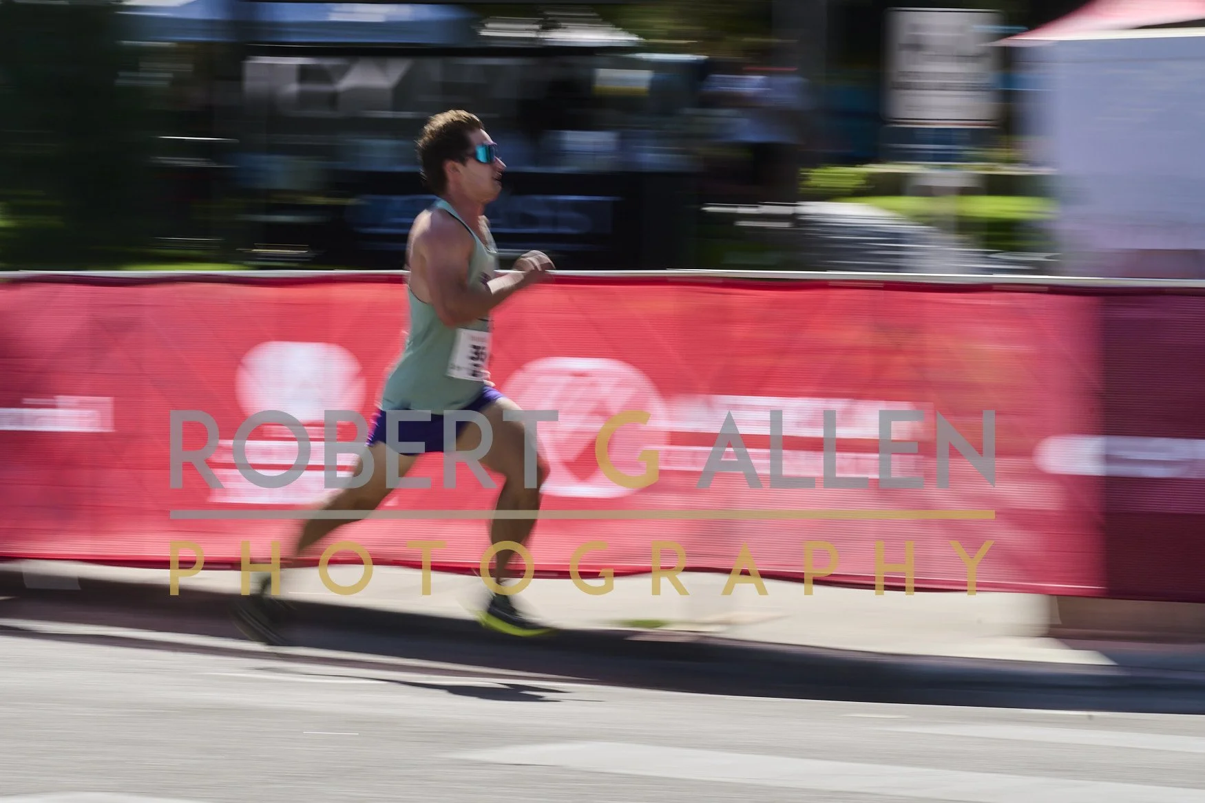 A male runner in a sleeveless shirt and shorts running past a pink banner with 'Robert Gallen Photography' text, with blurred background indicating motion.