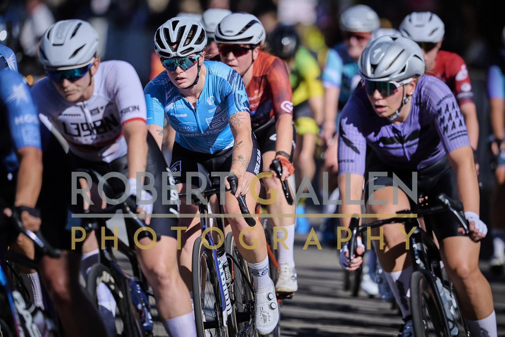 Close-up of female cyclists in a race wearing helmets and sunglasses, riding on a road with a crowd in the background.