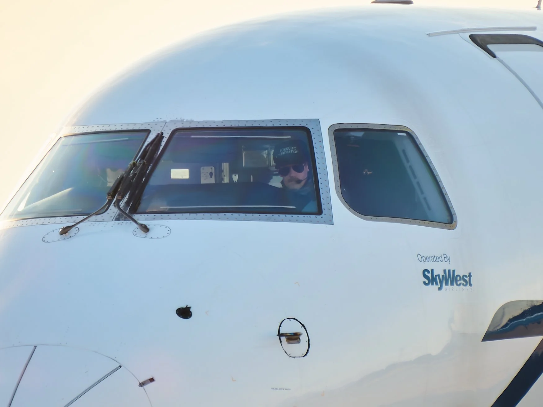Close-up of airplane cockpit with a pilot wearing sunglasses, and the logos of SkyWest Airlines visible on the aircraft fuselage.