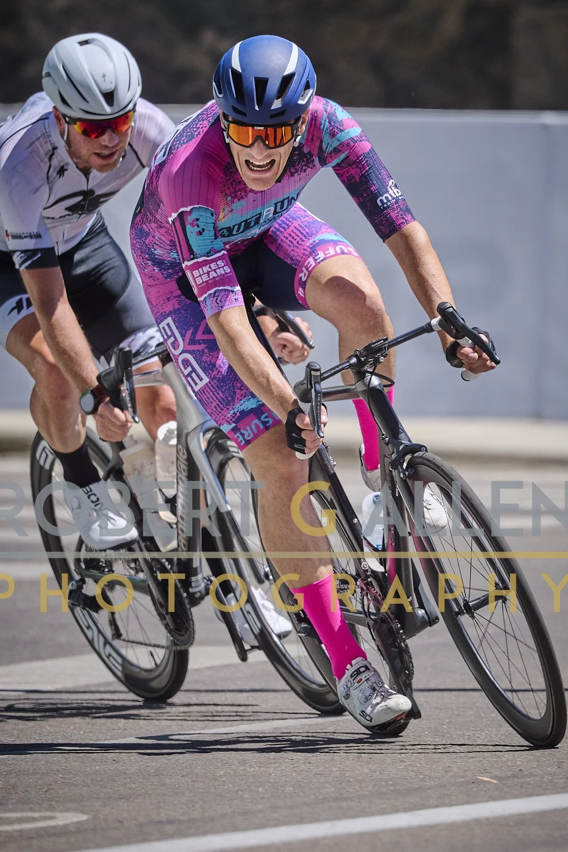Two cyclists riding on a road. The cyclist in the front is wearing a pink and blue cycling outfit, a blue helmet, and sunglasses. The cyclist behind is wearing a white outfit and a white helmet. They appear to be racing or training, leaning into a tu