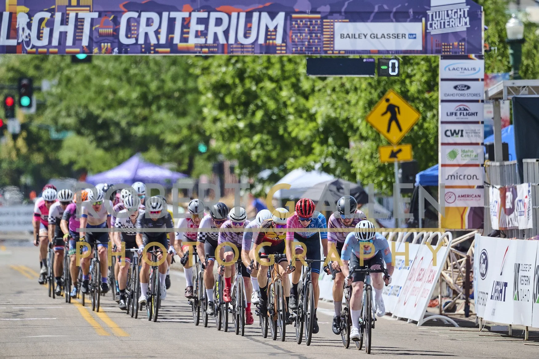 Cyclists racing in a group during a competitive cycling event, with a banner overhead and spectators in the background.