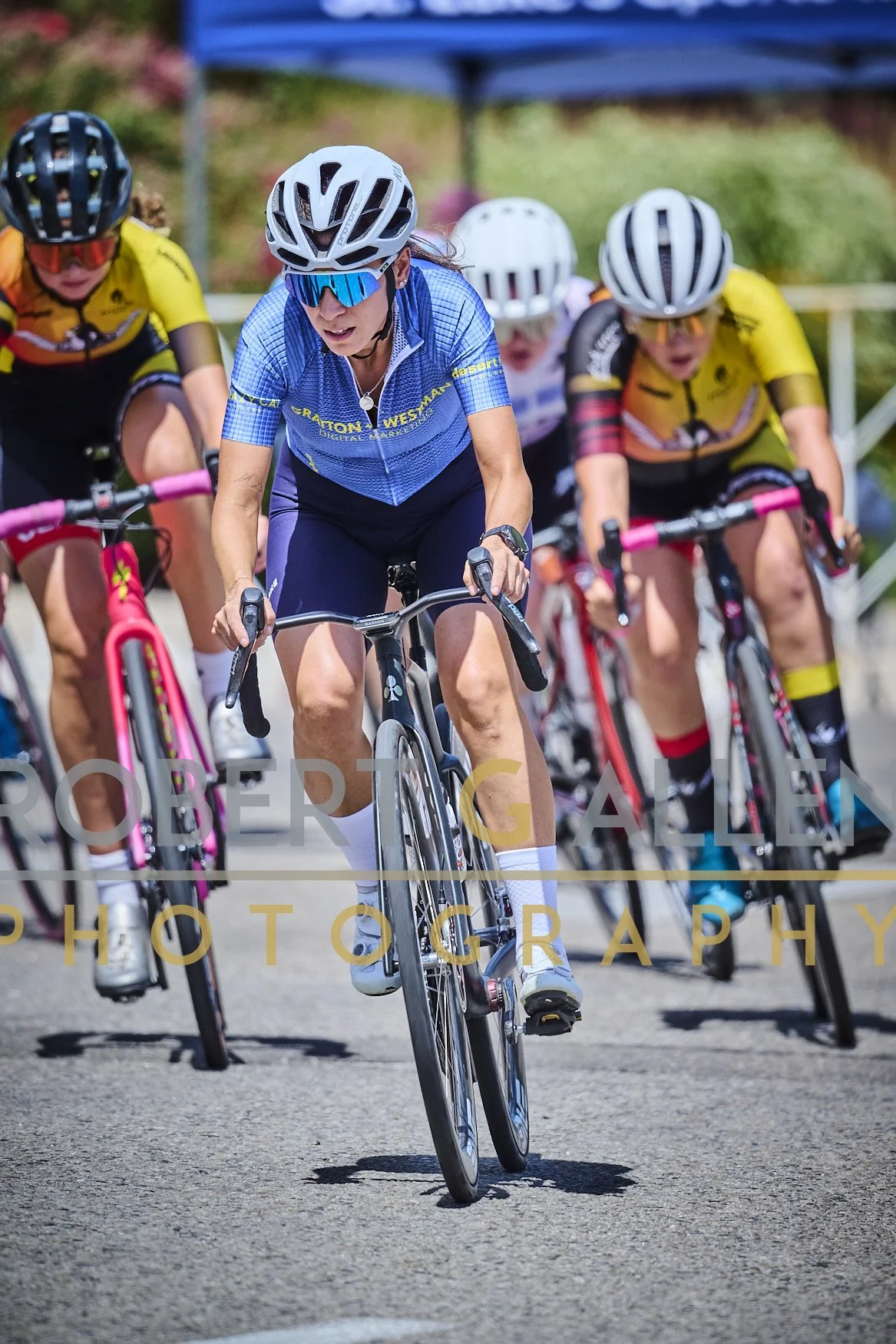 A group of female cyclists participating in a race, with three women visible in the foreground wearing helmets and sunglasses, riding on a paved road.
