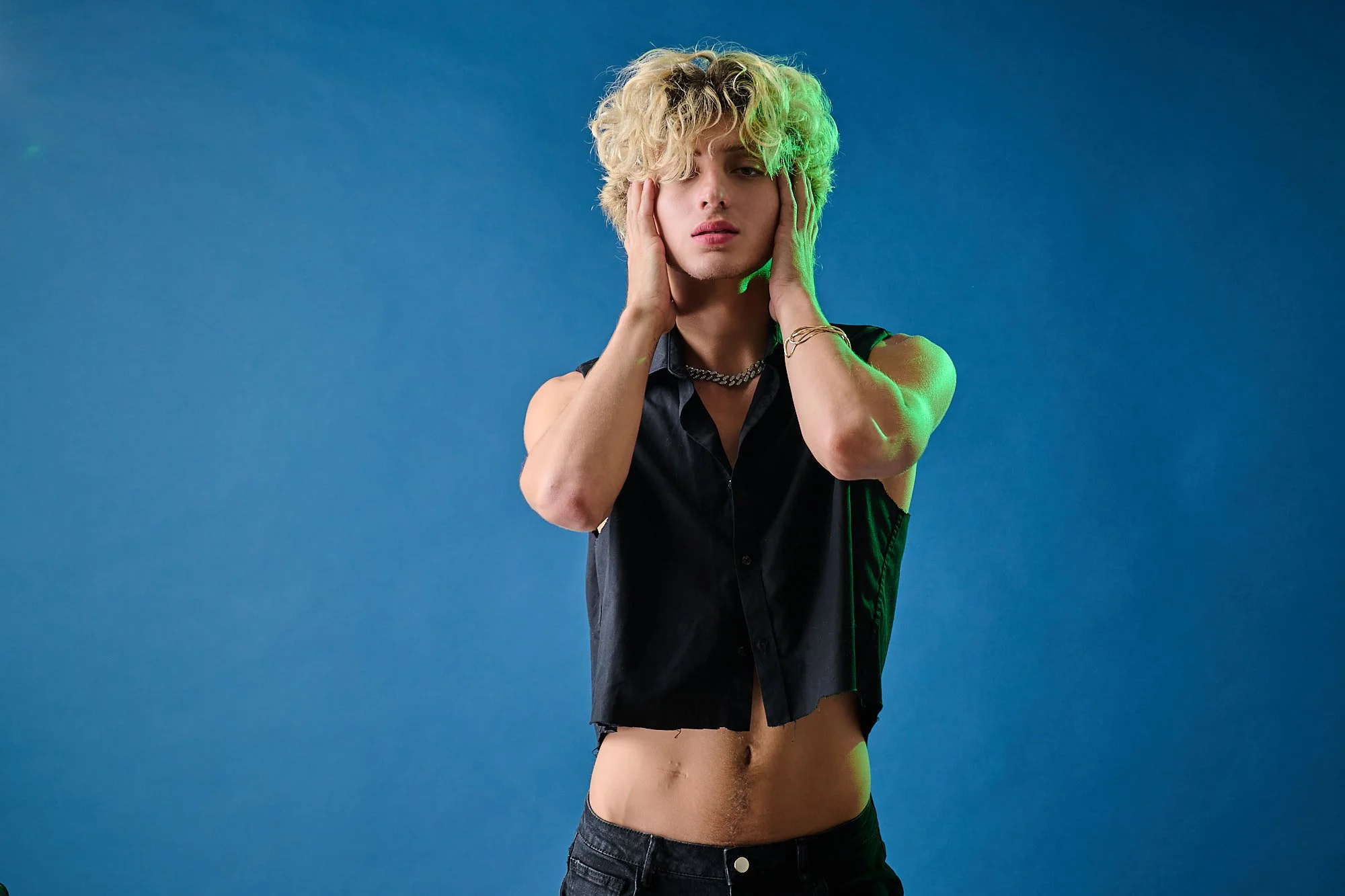A young person with curly blond hair posing with hands on face, wearing a sleeveless black shirt and jeans against a blue background.