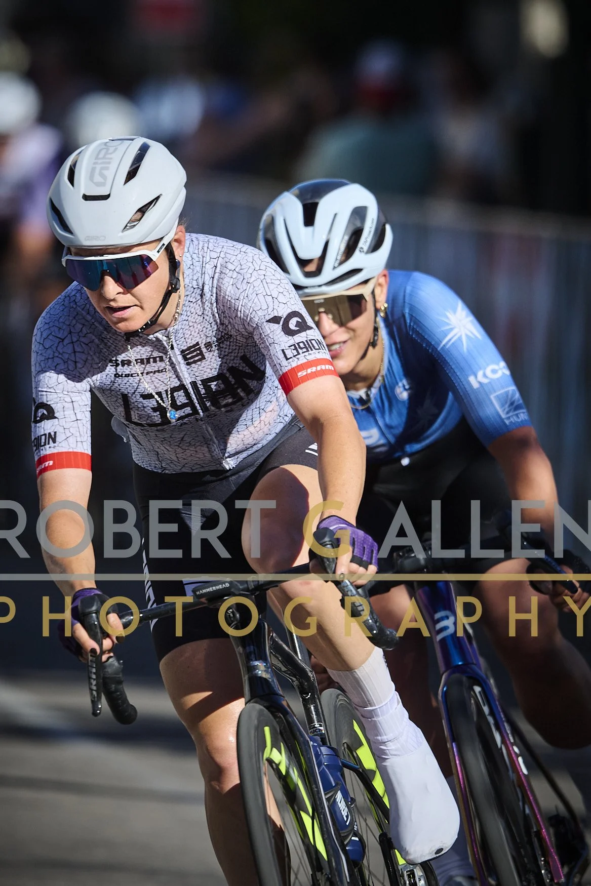 Two women cycling closely together during a race, wearing helmets, sunglasses, and team jerseys, with a blurred background of spectators.