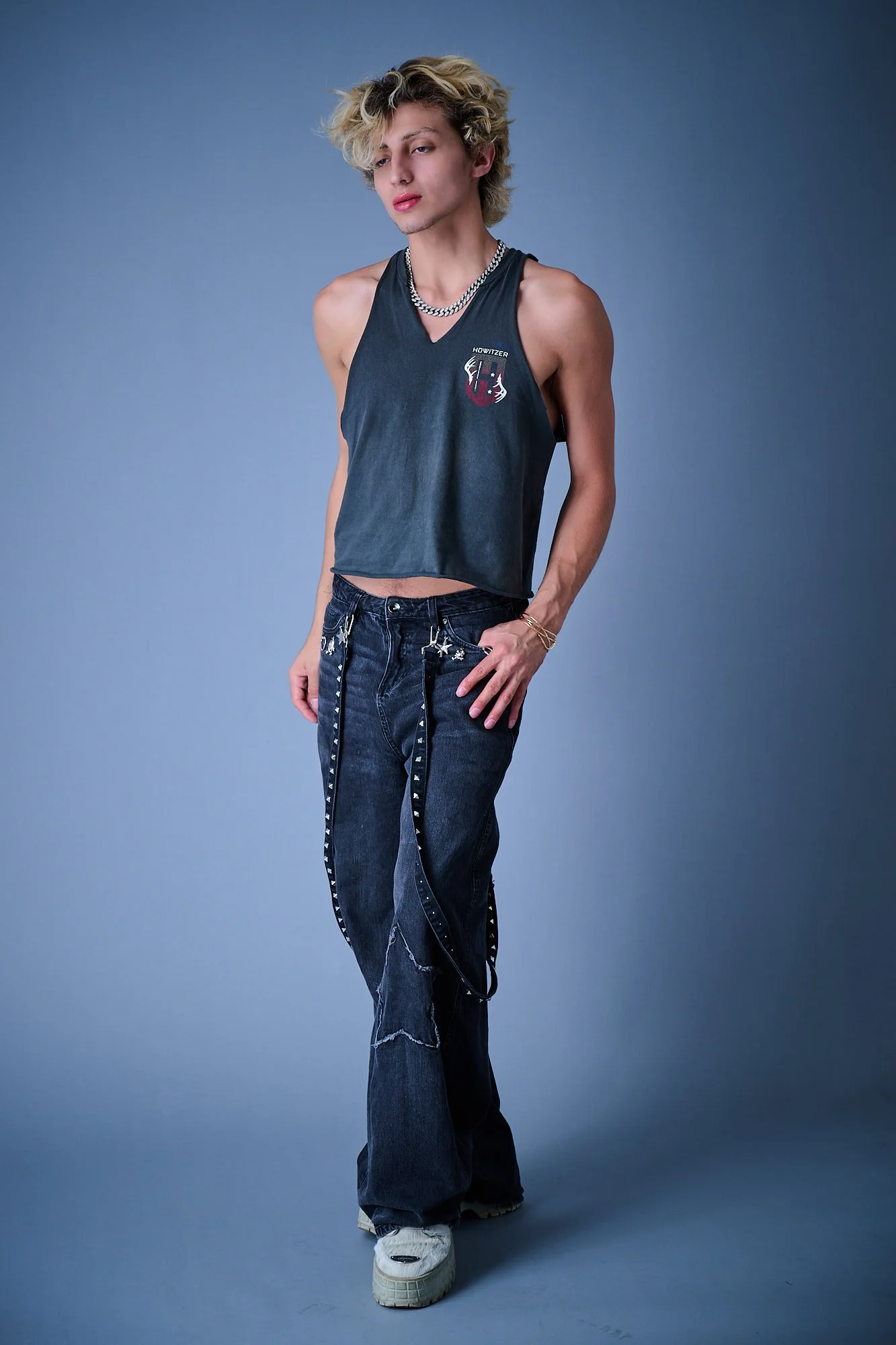 A person with light skin and styled blonde hair poses against a blue backdrop, wearing a black sleeveless top, distressed jeans with chains, white sneakers, a chunky silver chain necklace, and gold jewelry, with a confident expression.