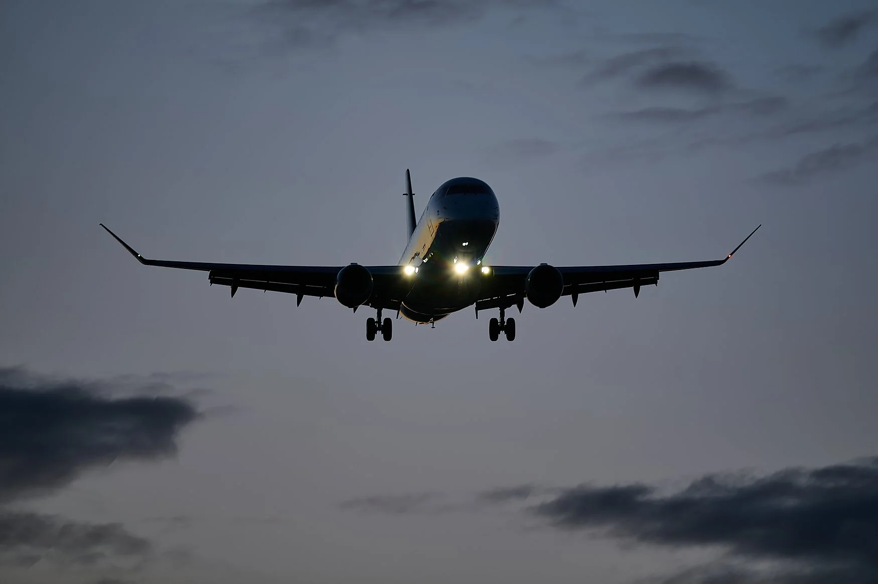 An airplane flying low in the evening sky with its lights on, surrounded by clouds.