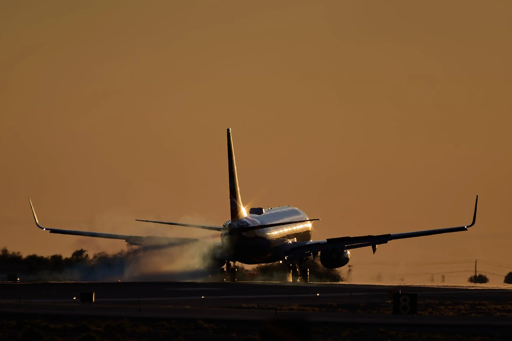 A commercial airplane on the runway at sunset, with sunlight reflecting off its body and tail, and dust or smoke around the landing gear.