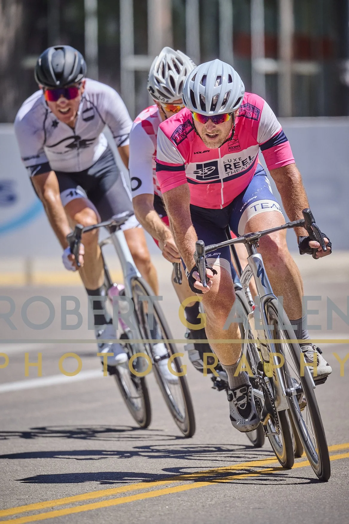 Three cyclists participating in a race on a paved road, wearing helmets and colorful jerseys, leaning forward and pedaling intensely.