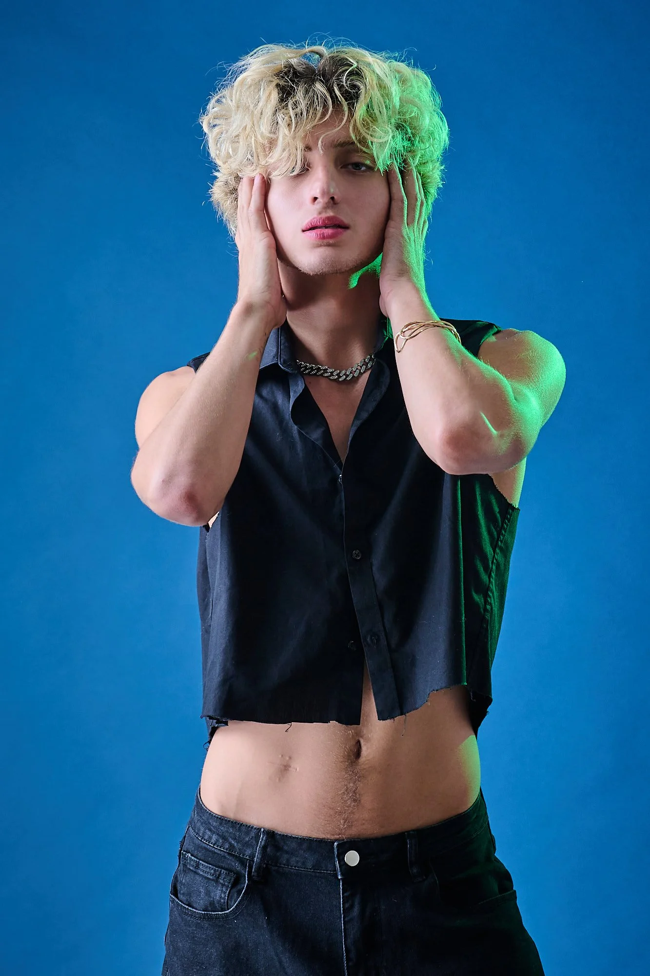 A young person with blonde, curly hair covering one eye, wearing a sleeveless black shirt, necklaces, and bracelets, posing with hands on their face against a blue background.