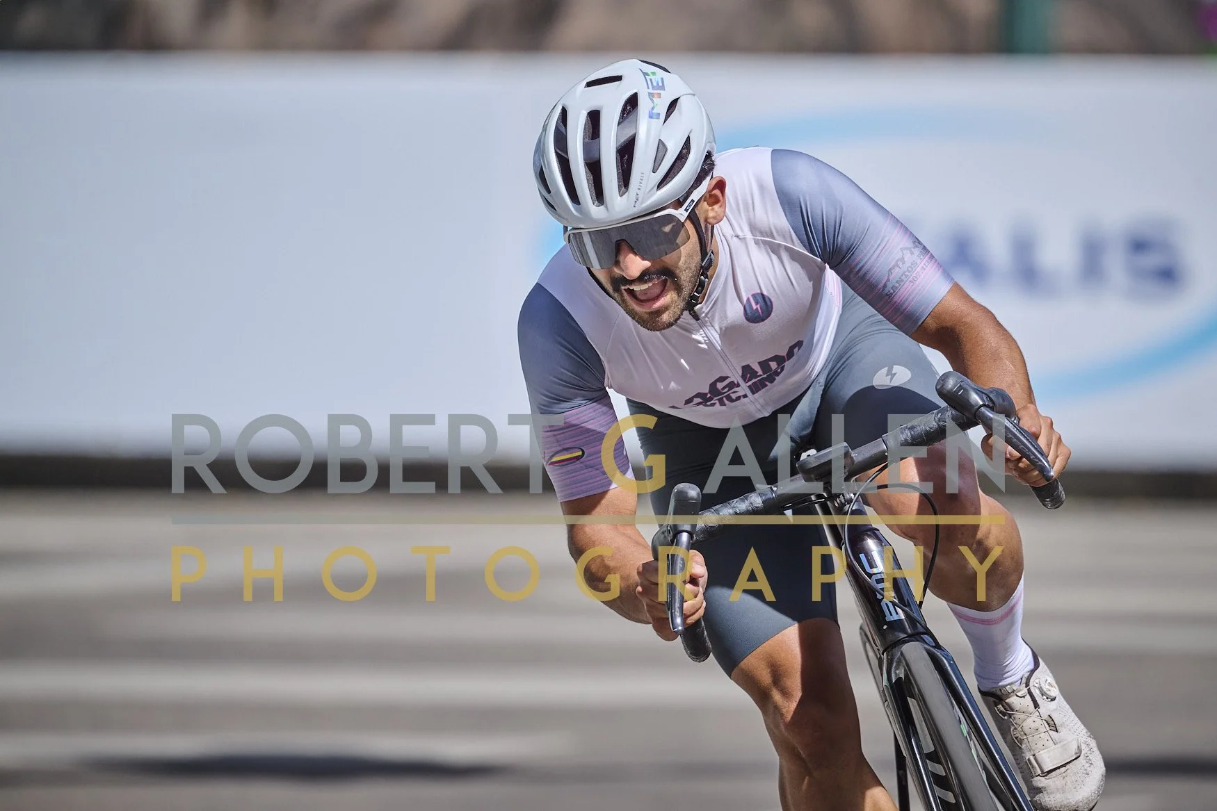 A cyclist wearing a white helmet, sunglasses, and a gray and white cycling jersey, riding speedily on a road with a blurred background.