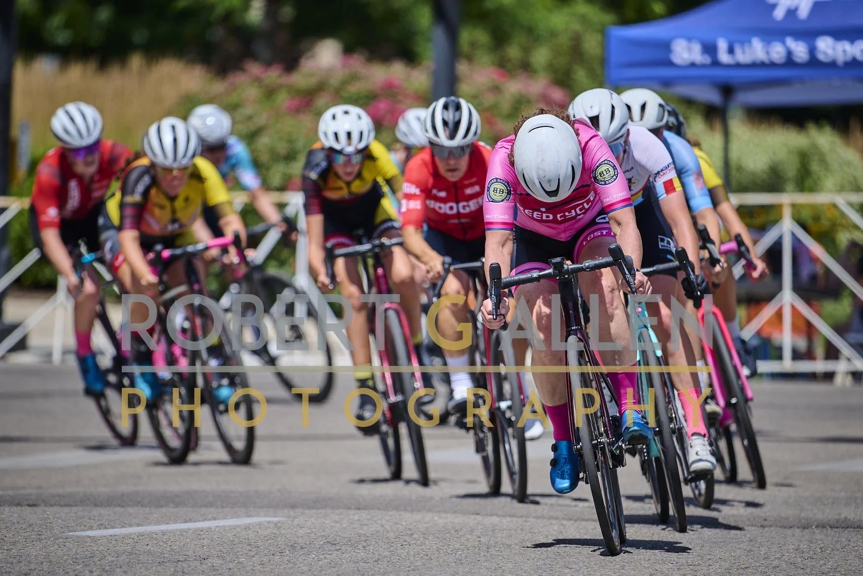 A group of female cyclists wearing helmets and colorful athletic jerseys racing in a competitive cycling event on a street, with some spectators and event tents in the background.