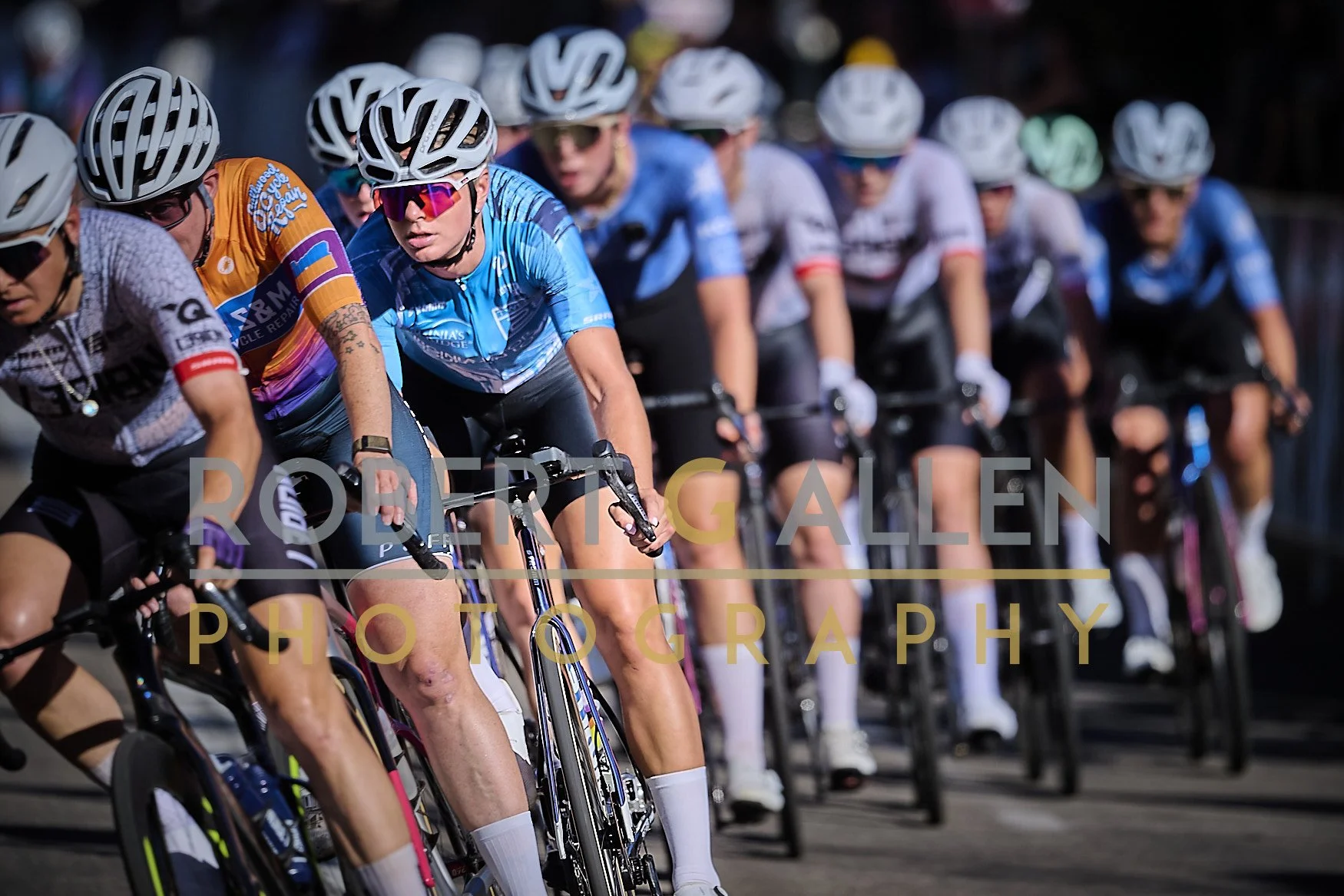 A group of female cyclists competing in a race, wearing helmets and team jerseys, riding closely together.