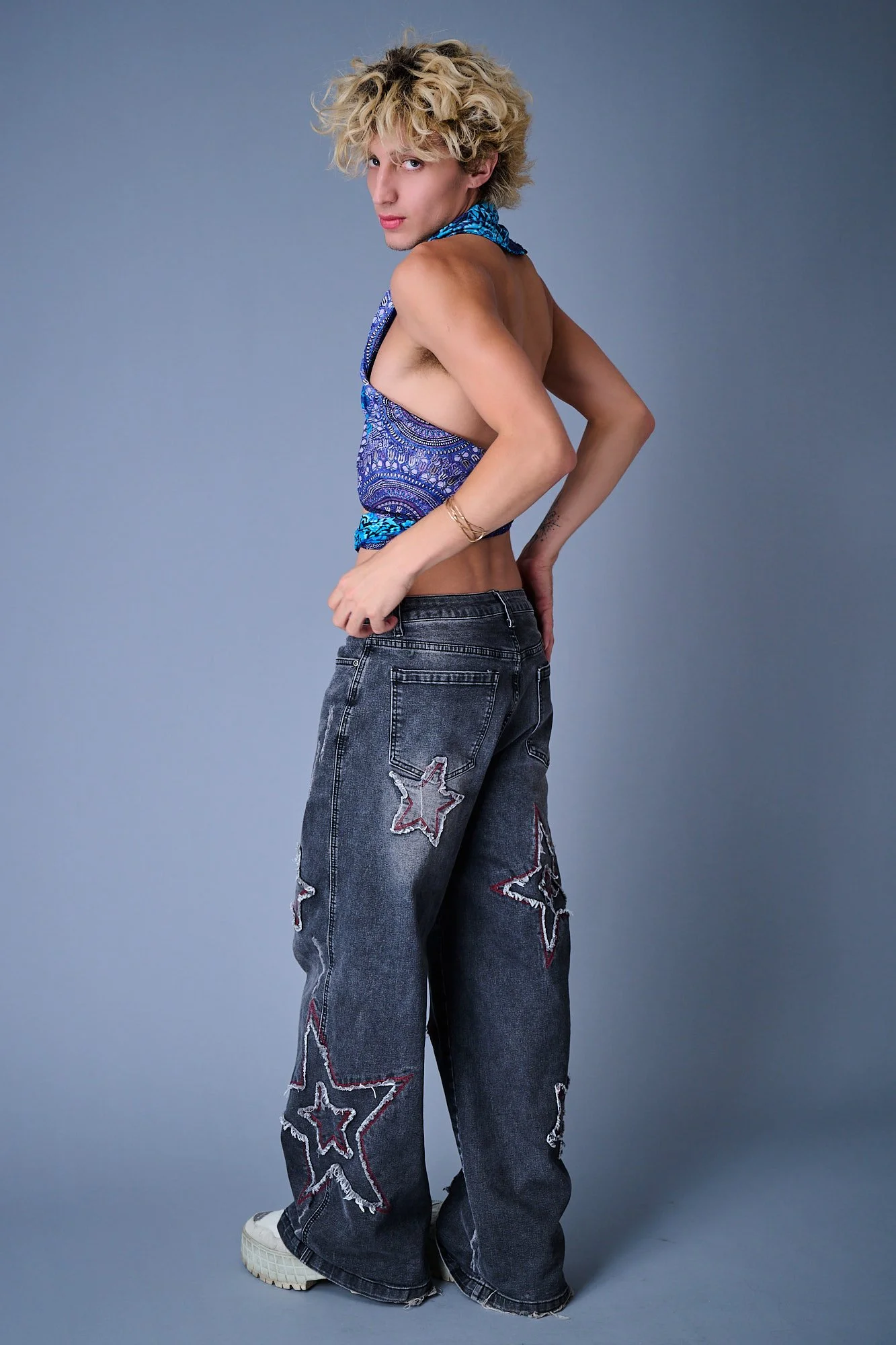 A young person with curly blonde hair, wearing a sleeveless blue patterned top, baggy jeans with star patches, and white sneakers, standing against a plain gray background and looking at the camera.