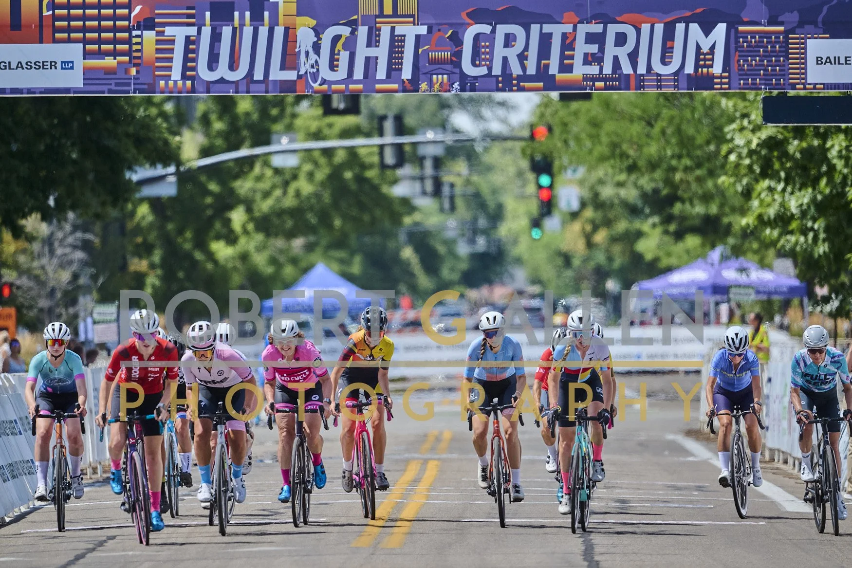 Cyclists participating in a race crossing a street under a colorful banner that reads 'Twilight Crit' with trees and setup tents in the background.