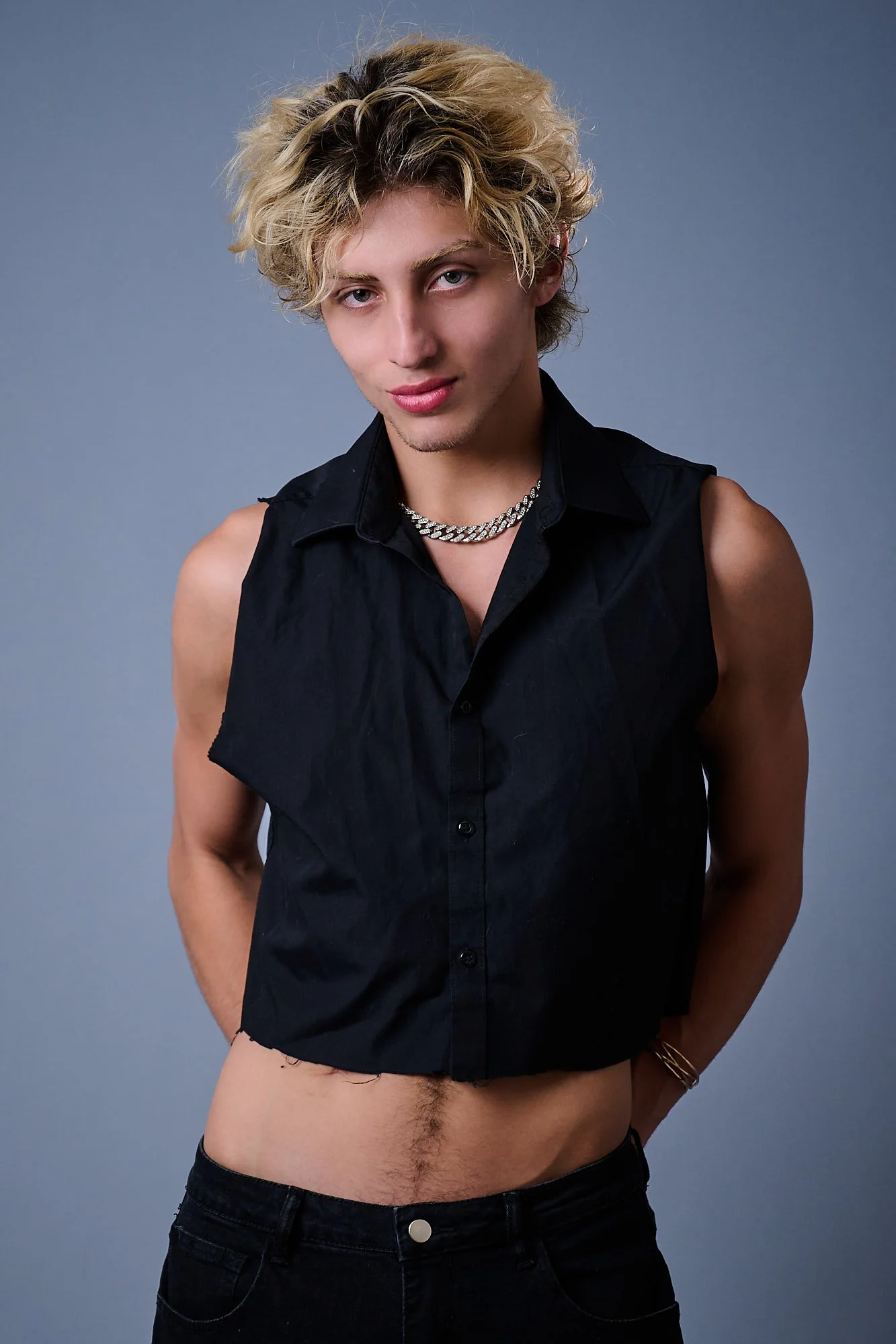 A young man with blonde, curly hair wearing a black sleeveless shirt, a silver chain necklace, and dark jeans, standing against a plain gray background.