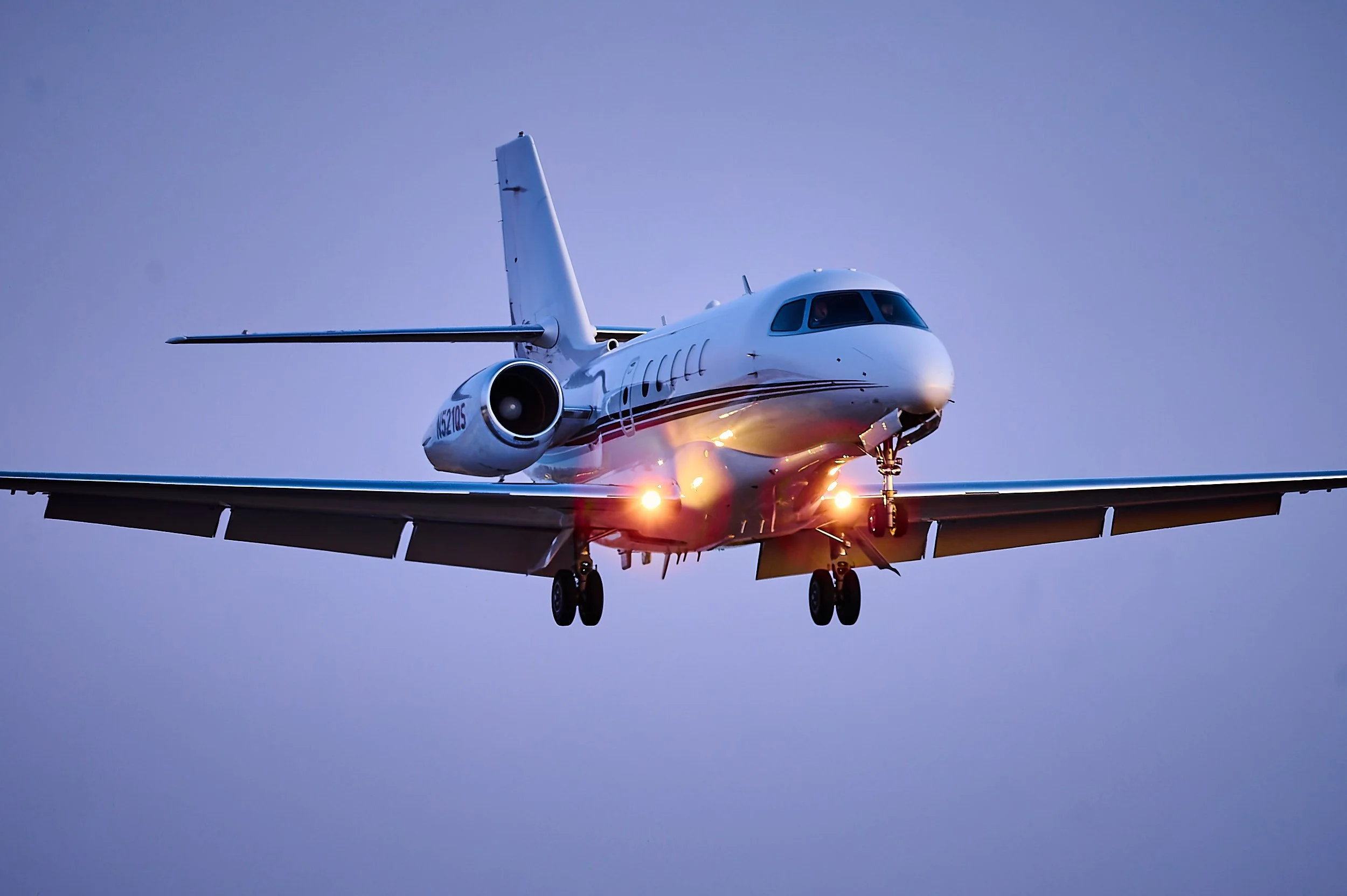 A white private jet airplane with red and black stripes flying through the sky during dusk or dawn.