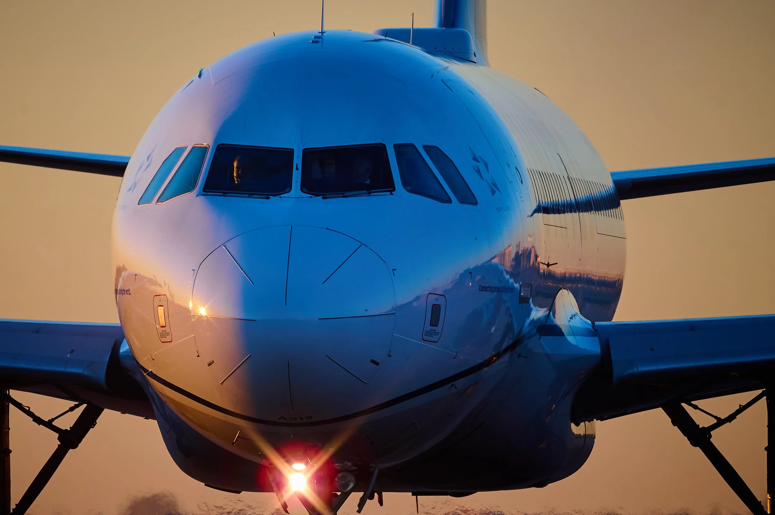 A close-up of a large commercial airplane's nose section with the cockpit windows visible, set against a colorful sunset sky.