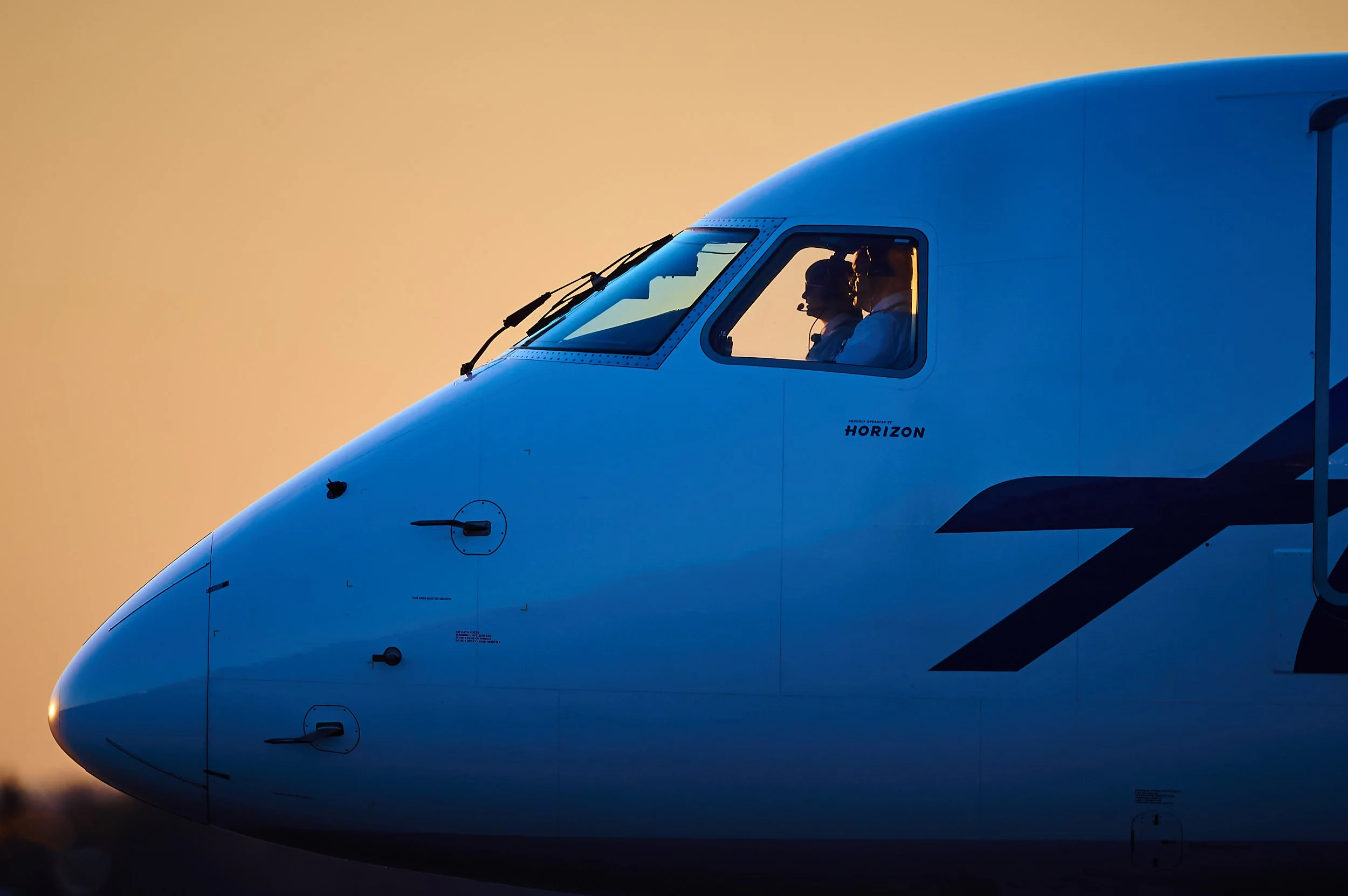 Close-up of the nose of a commercial airplane with two pilots visible through the cockpit window, set against a soft orange sky at sunset.