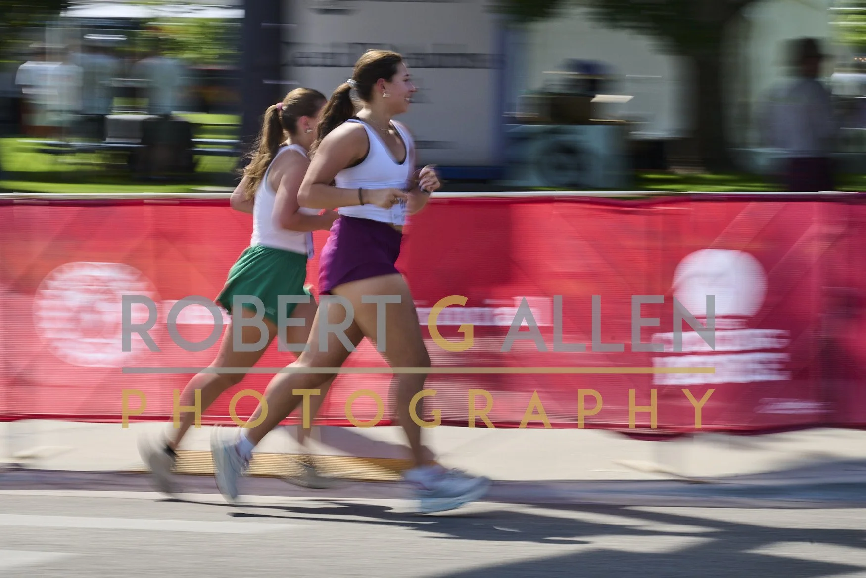Two female runners competing in a race, running past a red barrier with 'Robert Gallen Photography' written on it. The background shows blurred spectators and structures, indicating motion.