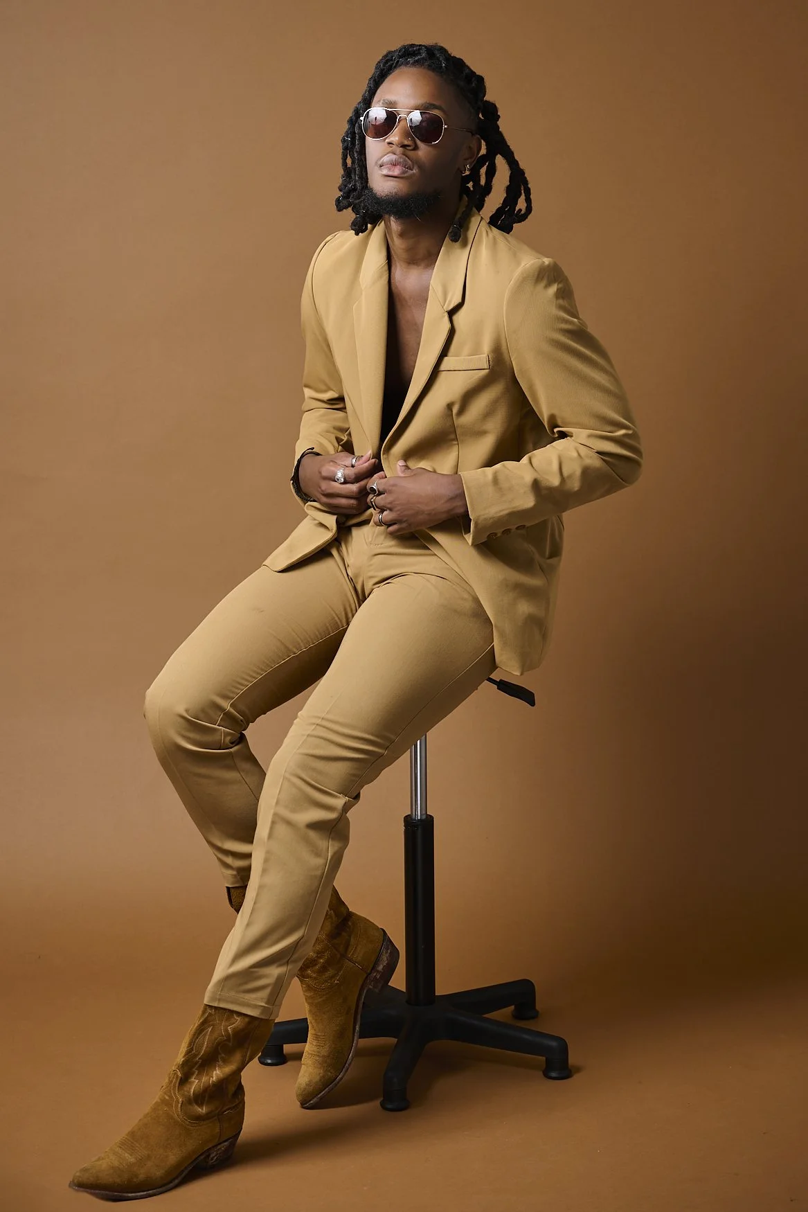 A man with dreadlocks, sunglasses, and a beard, wearing a tan suit and brown boots, seated on a stool against a plain tan background.