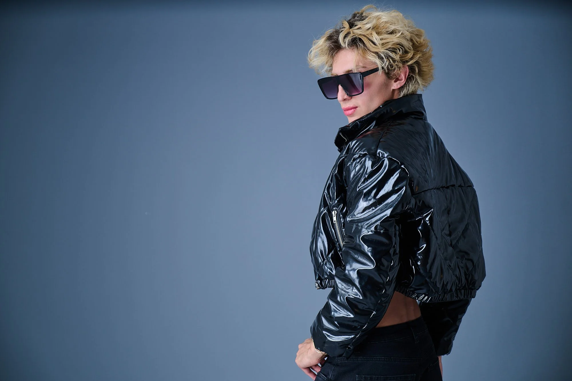 A person with short, curly blonde hair wearing large black sunglasses and a shiny black leather jacket. They are standing sideways against a plain gray background.