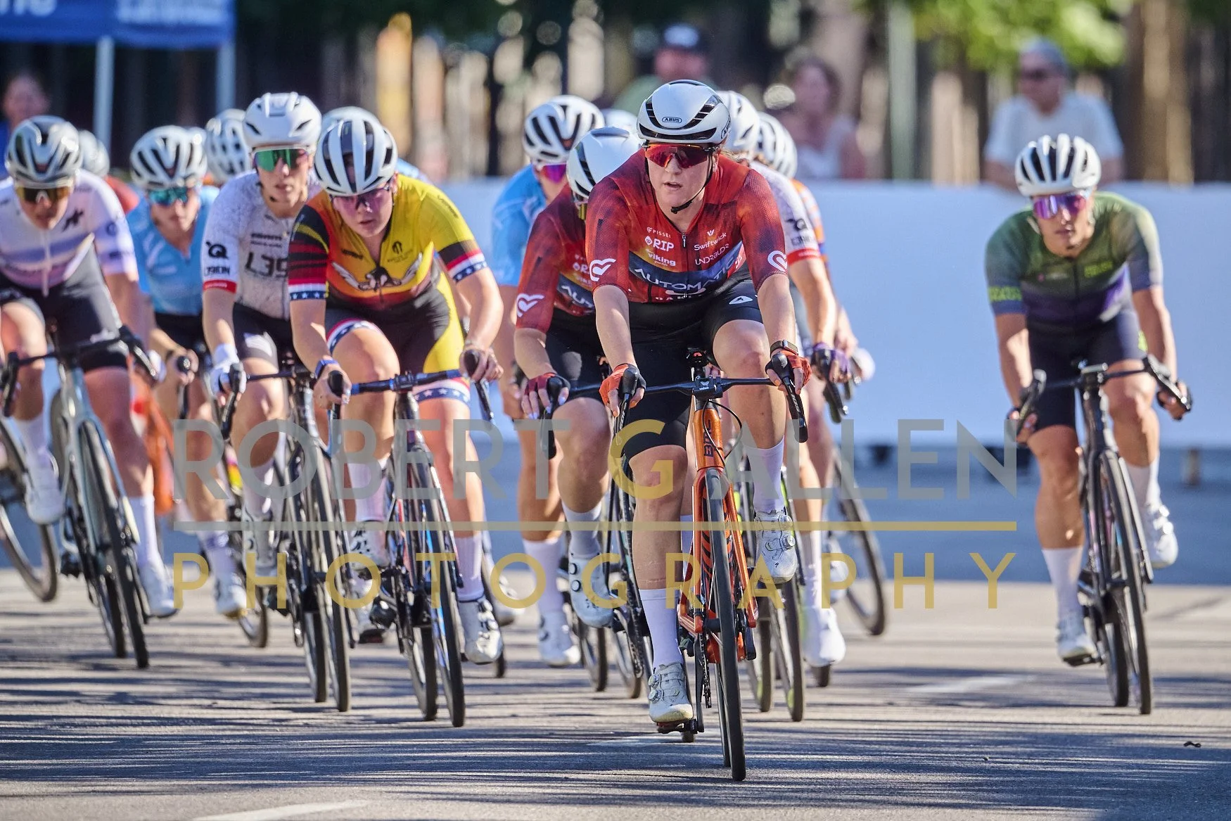 A group of female cyclists participating in a race, wearing helmets and sunglasses, riding bikes in close formation outdoors on a sunny day.