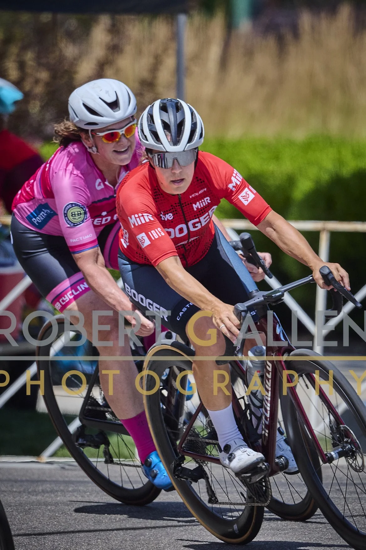 Two women riding bicycles during a race or event, both wearing helmets and sports sunglasses. The woman in front wears a red jersey with black shorts, and the woman behind wears a pink jersey with black shorts. They are leaning into a turn on a paved