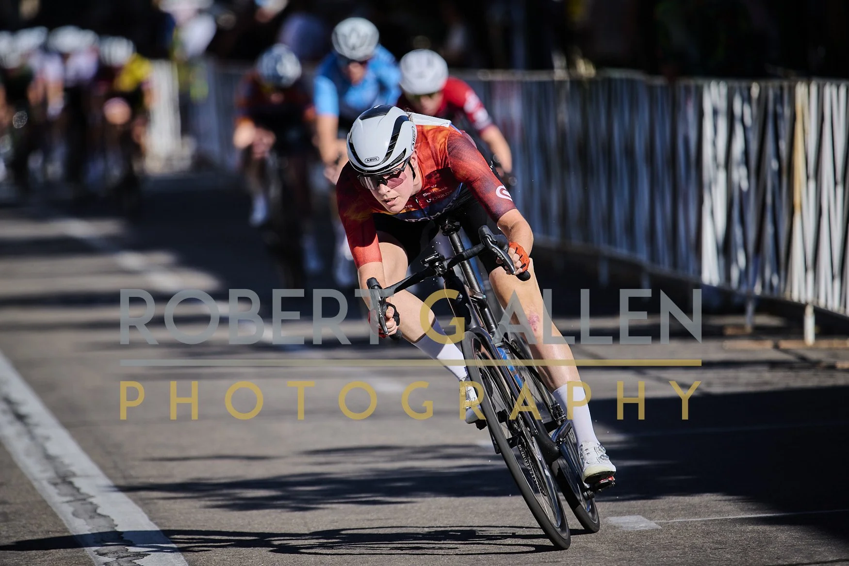 A cyclist wearing a white helmet and red and black outfit leans during a bicycle race on a paved street with metal barriers and several other racers in the background.
