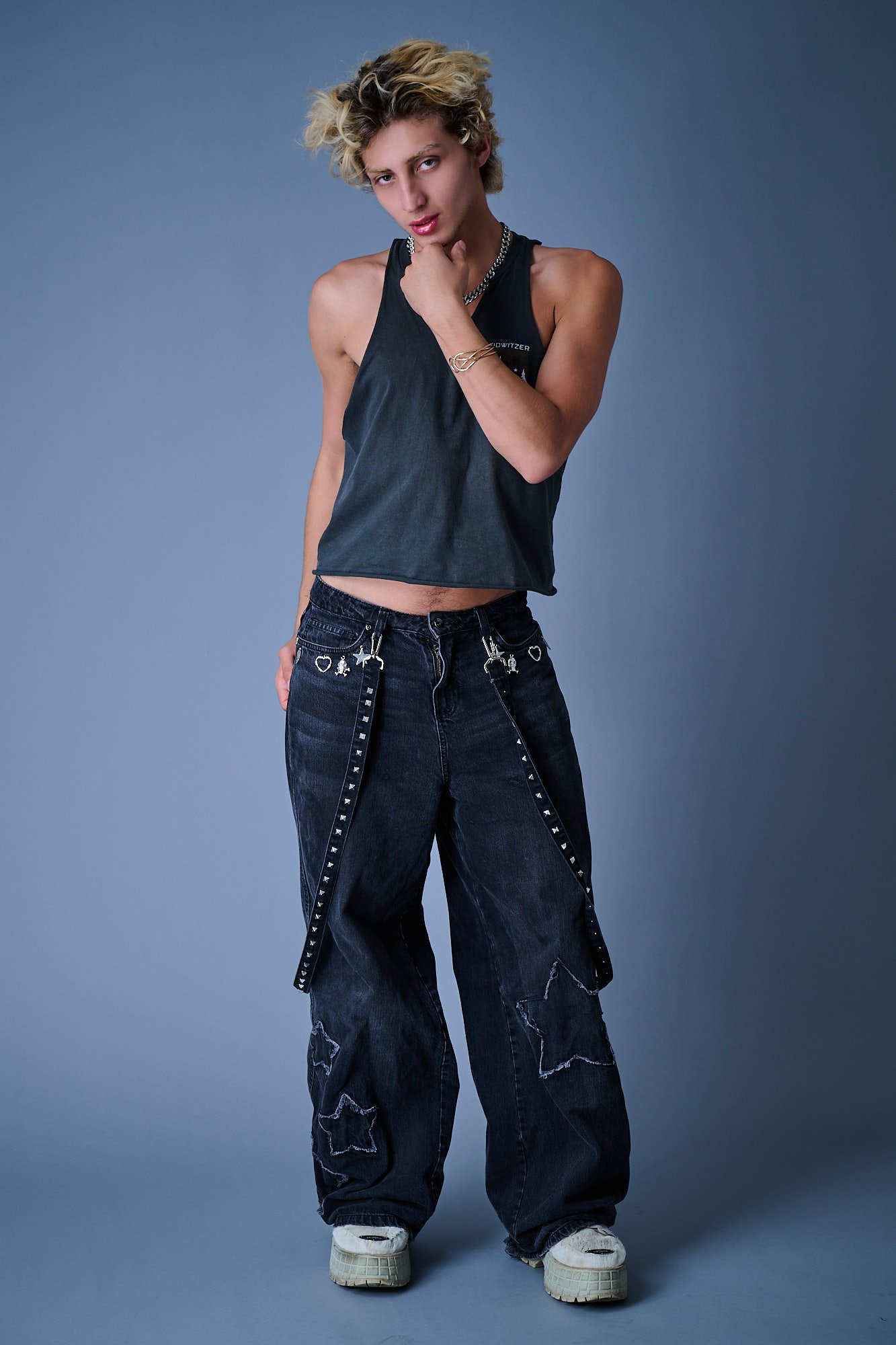 A young person with short, curly blonde hair, wearing a sleeveless black top, baggy ripped jeans with star and heart embellishments, and chunky white sneakers, standing against a plain blue background with one hand resting on their chin.