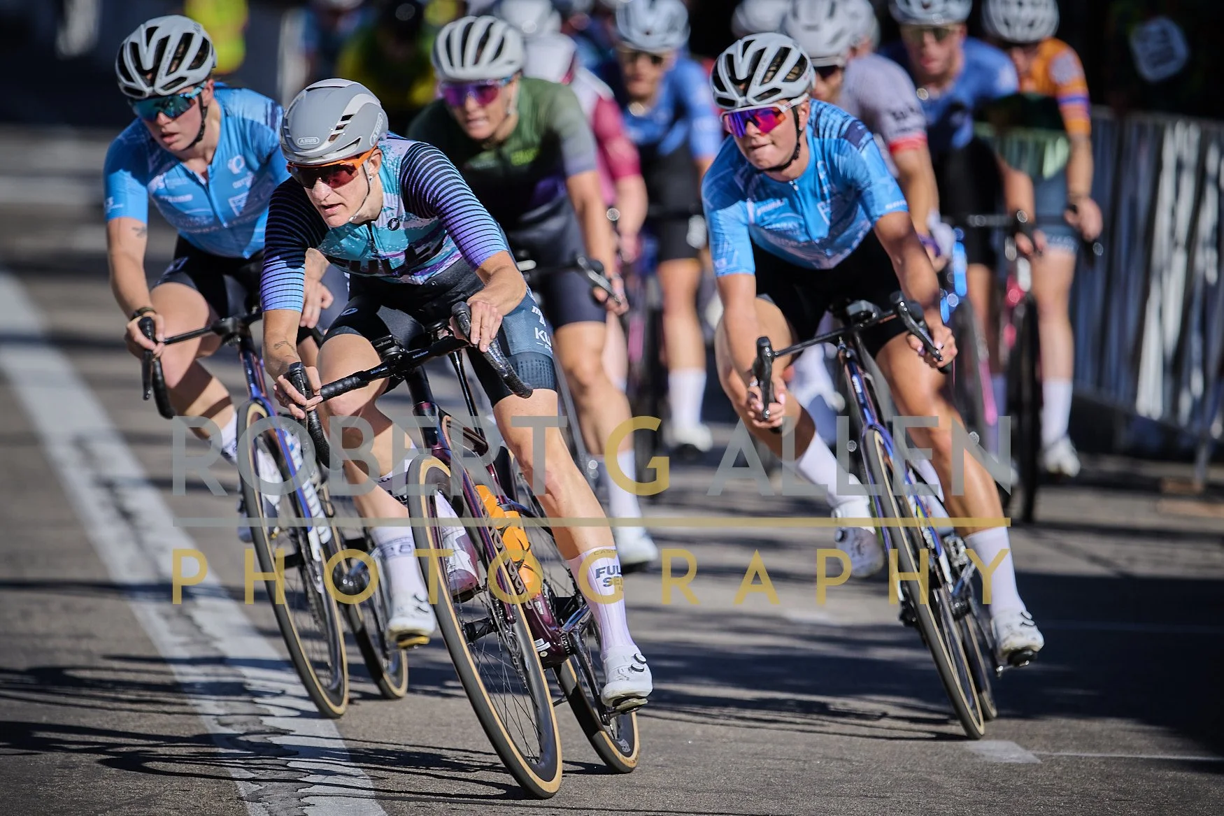Group of female cyclists racing on a velodrome track, dressed in colorful cycling jerseys and helmets, leaning into a turn.